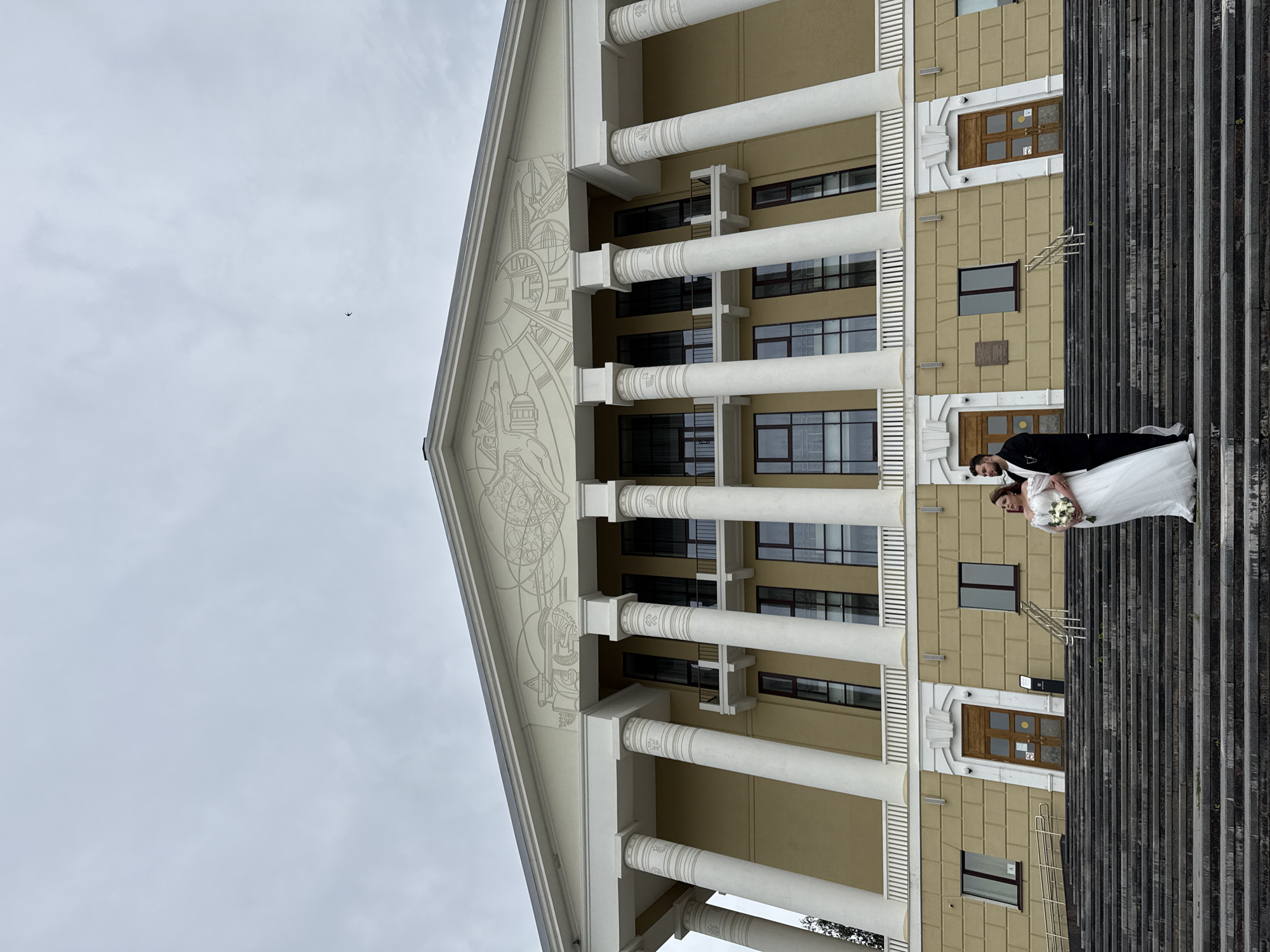 A wedding couple stands on the wide, stone steps leading up to a grand, yellow building with large white columns and a classical facade. The bride, in a white wedding dress and holding a bouquet, stands to the left, looking up and to her right. The groom, in a black suit, stands to her right, looking to his right. A small bird is visible in the overcast sky above the building. The building appears to be an administrative or public building, possibly a palace of culture or a government institution. The scene is likely set in the city of Votkinsk, Russia, given the architectural style common in Soviet-era public buildings.