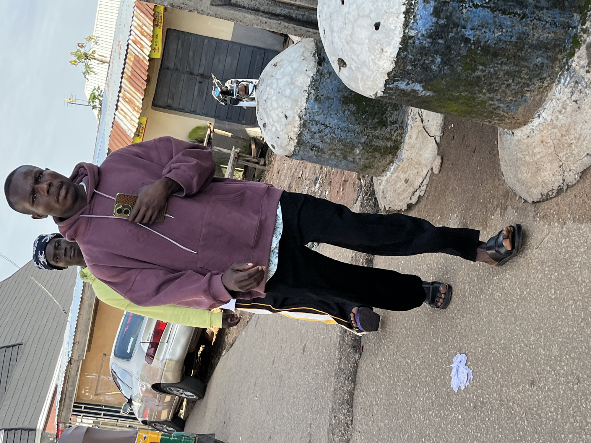 In the city of Jos, Nigeria, a group of individuals walk along a paved street with a car parked to the left and buildings in the background. A man in the foreground, wearing a maroon hoodie and black pants, holds a smartphone in his right hand. Another man in a lime-green hoodie walks behind him, and a third individual in a headscarf is partially visible behind them. Two large, rounded concrete structures, possibly bollards, are in the right foreground, showing signs of wear and moss growth. A motorcycle is parked near a doorway in the background. The scene appears to be an outdoor street setting.
