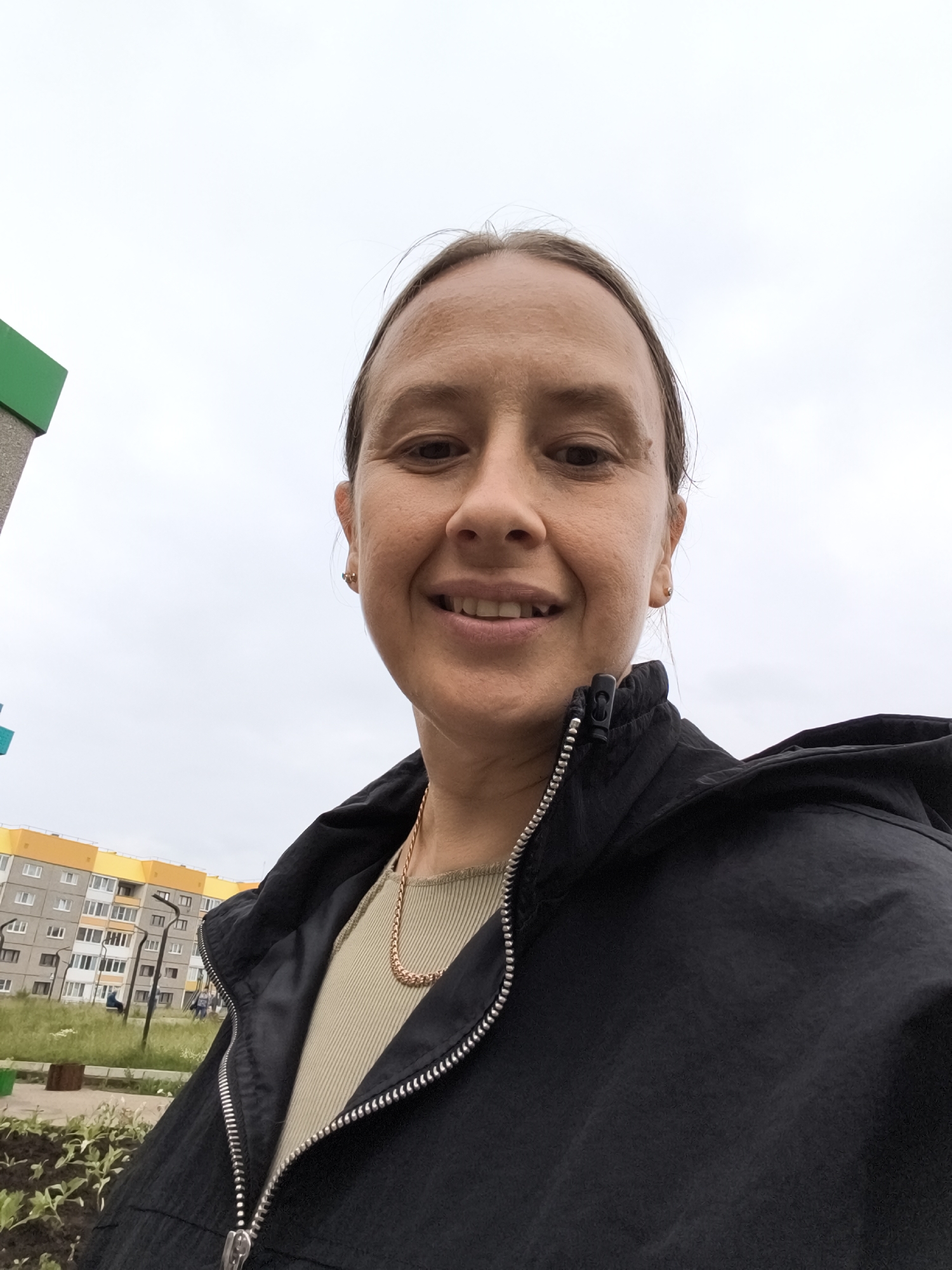 A medium shot, outdoor selfie depicts a fair-skinned woman with medium-length brown hair, pulled back, smiling as she looks directly at the camera. She is wearing a light olive green, ribbed, short-sleeved shirt, a gold chain necklace, and a black zippered jacket. The background features a cloudy sky, a section of a gray and green structure on the left, and a low-rise apartment building with yellow accents in the distance. The apartment building is located in the residential area of Марковский, Russia, a district within the city of Ulan-Ude. The immediate foreground shows a small patch of disturbed earth with some green seedlings.