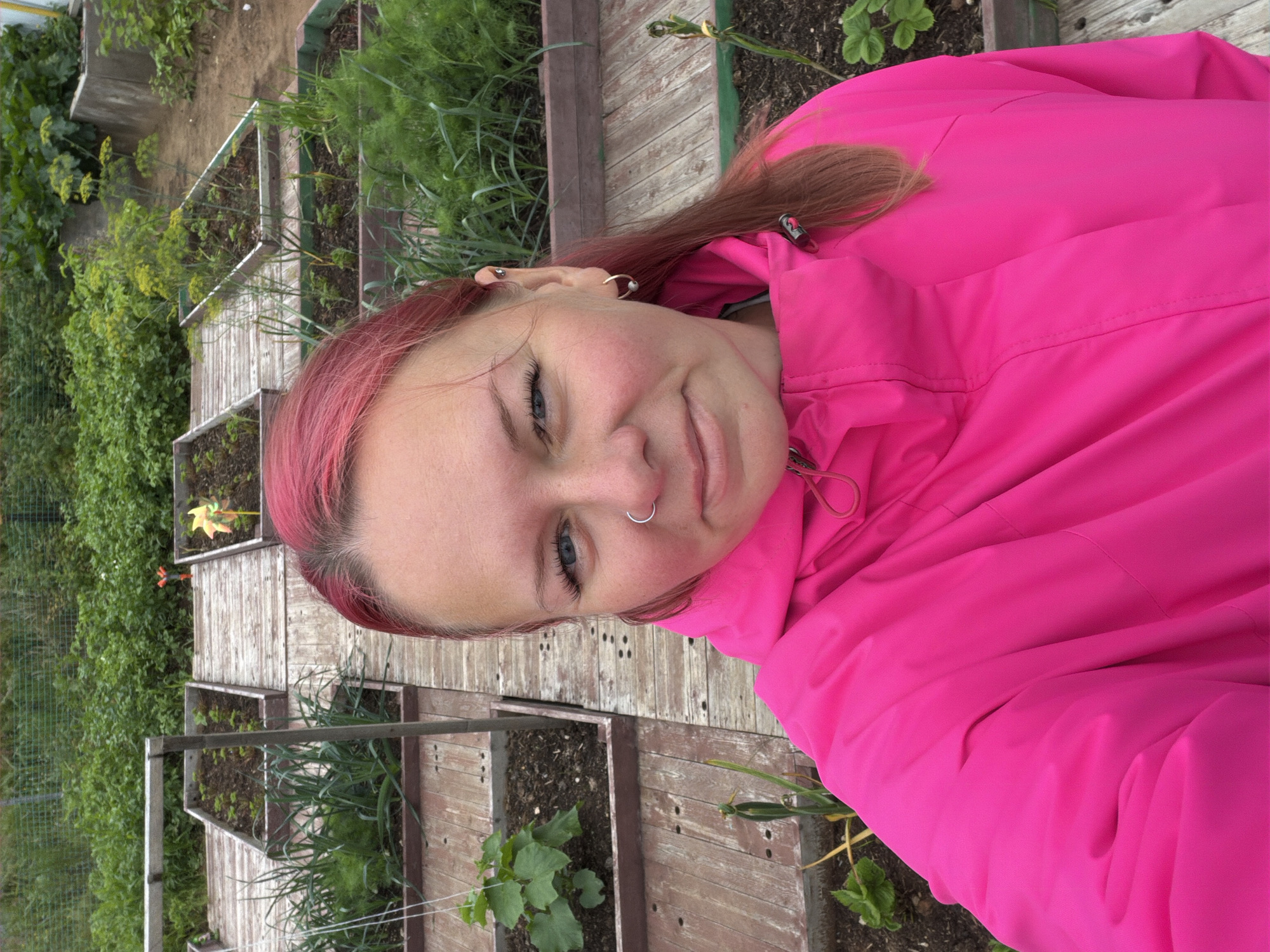 A woman with bright pink hair and a pink jacket is taking a selfie in a garden. Raised garden beds are visible in the background, some containing plants like leeks and dill. The garden beds are constructed with wooden planks. A small, colorful pinwheel is planted in one of the beds. The ground between the beds is also made of wooden planks. The overall environment appears to be an outdoor garden space. The image was likely taken in Chaikovsky, Russia, given the context.