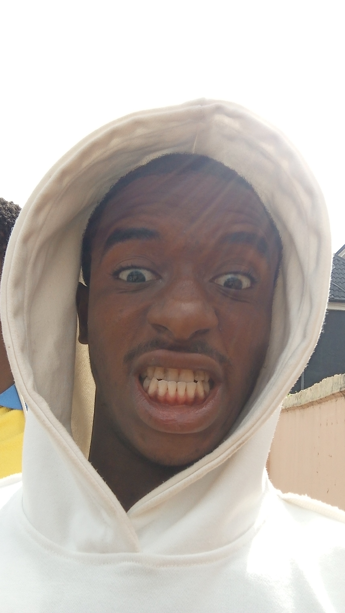 A close-up, full-frontal shot reveals a dark-skinned man with a thin mustache and wide, slightly crossed eyes, wearing a white hoodie. He is grinning broadly, displaying his upper and lower teeth. The hood of the hoodie frames his face. The background is blurry, revealing a light-colored wall and a partially visible building or structure. The presence of sunlight suggests an outdoor setting, and based on the prompt, the location is Obosi, Nigeria. There is another person partially visible on the left of the image.
