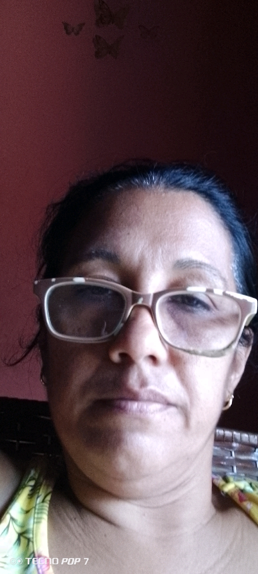 A medium shot selfie shows a woman wearing eyeglasses. She is looking directly at the camera, and her face fills most of the frame. She has dark hair pulled back, and her skin tone is medium to dark. The background is a dark red wall with a subtle butterfly decal. The lighting is somewhat dim, creating shadows on her face. There is a logo "TECNO POP 7" in the bottom left corner of the image. The image does not provide any explicit visual cues to determine the specific location within the city of Altagracia, Venezuela.