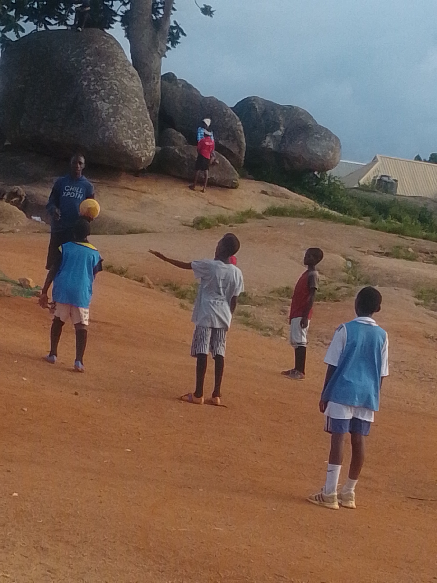 In an outdoor setting, a group of children appears to be playing with a ball. The environment is characterized by a dirt ground and large rock formations. A tall tree is visible on the left side of the image. In the background, a building with a yellow roof can be seen. There are at least five children in the scene, some are actively engaged with the ball, while others appear to be observing. The lighting suggests it could be late afternoon or early morning.