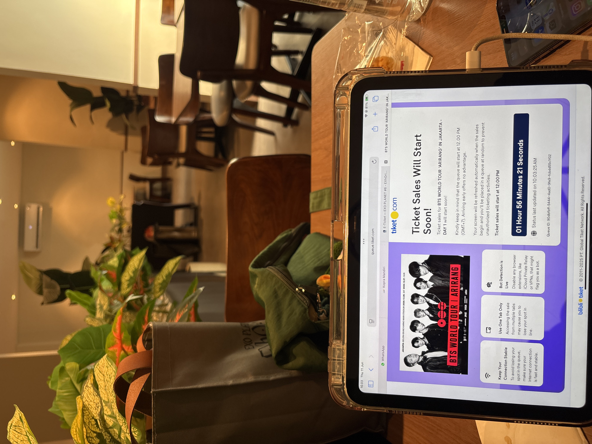An indoor shot shows a tablet displaying a ticket sale announcement for "BTS WORLD TOUR 'ARIRANG' IN JAKARTA - DAY 1". The tablet is placed on a wooden table, with a charging cable connected to a smartphone resting beside it. In the background, a cafe or restaurant setting is visible with wooden chairs and tables, and potted plants with lush green and reddish-orange leaves. The time on the tablet's status bar indicates 10:03 Thu 11 Jun. The ticket sale is for an event at JAKARTA GELORA BUNG KARNO MAIN STADIUM on 2026.12.26 and 2026.12.27. The announcement highlights that ticket sales will start soon, with the queue starting at 12:00 PM (GMT+7). A countdown timer displays "01 Hour 56 Minutes 21 Seconds" for the status update at 10:03:25 AM. The image appears to be taken at a venue in West Jakarta, Indonesia, given the event's location.