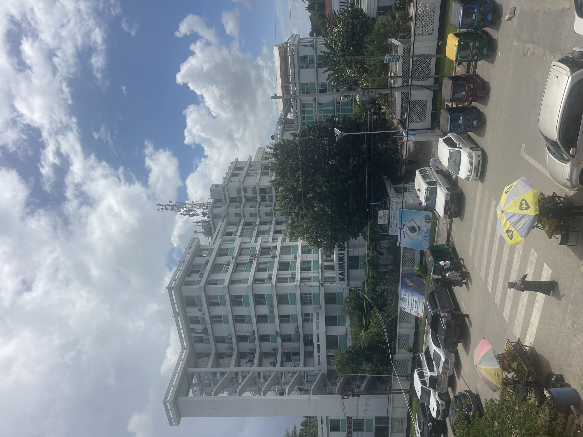 This image captures a vibrant street scene in Dar es-Salaam, Tanzania, under a bright, partly cloudy sky. The central focus is a large, multi-story white building with distinct green or teal window accents and numerous balconies, resembling fire escapes, on its facade. The building's name, "KAIRUKI HO," is visible on its side, and a nearby sign explicitly states, "WELCOME TO KAIRUKI HOSPITAL," indicating its function. Communication antennas crown the top of this building.
The street below is a hive of activity. A diverse array of vehicles is present, including several cars (white, silver, and dark-colored) and multiple three-wheeled tuk-tuks, distinguished by their colorful tops (blue, green/yellow, red, and dark blue). A white ambulance is also part of the street traffic. Several individuals are seen walking on the sidewalks and street, including one person in dark clothing crossing a zebra-striped pedestrian crosswalk.
A street vendor is visible on the right, operating from a cart filled with fruits, shaded by a distinctive yellow and white umbrella marked with "SDH." Another smaller, multi-colored umbrella (pink and yellow) is also present. The urban environment includes streetlights, power lines, and dense green foliage from trees. The time of day is clearly daytime, with bright sunlight casting visible shadows, and the weather appears fair with scattered white clouds against a blue sky. The scene depicts a typical bustling urban environment in a tropical setting.