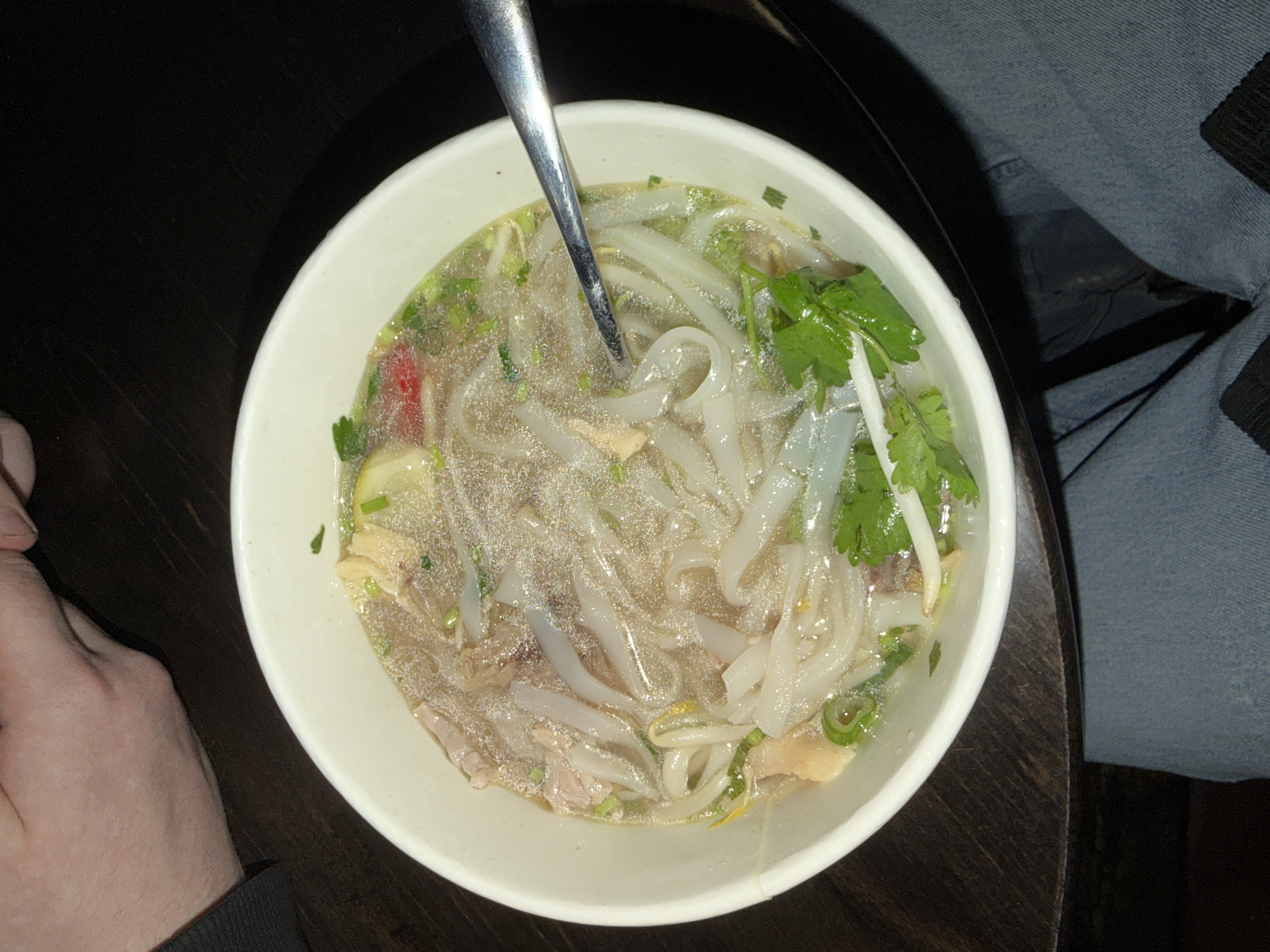 A high-angle close-up shot shows a white bowl filled with Vietnamese pho soup. A metal spoon rests inside the bowl, partially submerged in the broth, noodles, and other ingredients, including green herbs. The bowl sits on a dark wooden surface. A person's hand and the cuff of a dark gray sleeve are visible on the left side of the frame. On the right side, part of a person's leg clad in blue denim fabric is visible. The location cannot be determined from the image.