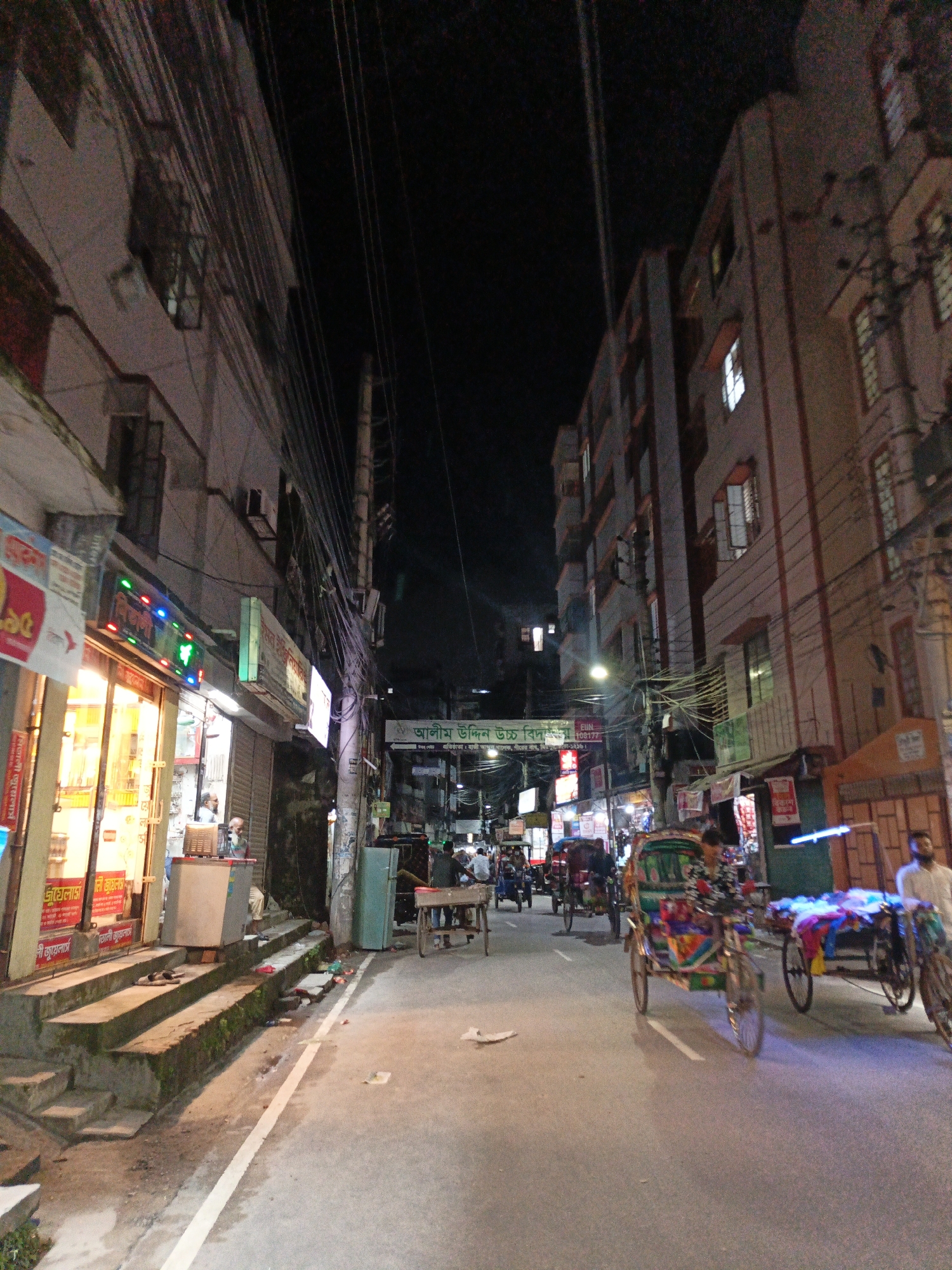 The image depicts a nighttime street scene in Dhaka, Bangladesh. The street is lined with multi-story buildings with illuminated windows and storefronts. A prominent banner with Bengali text is visible across the street, suggesting a local establishment or event. The street is busy with various forms of transportation, including rickshaws, some of which are decorated with colorful fabrics. People are seen walking and interacting in the dimly lit environment. Overhead, a dense network of electrical wires crisscrosses the scene, a common characteristic of urban infrastructure in Dhaka. The overall atmosphere is that of a bustling city at night, with a mix of commercial activity and pedestrian traffic.