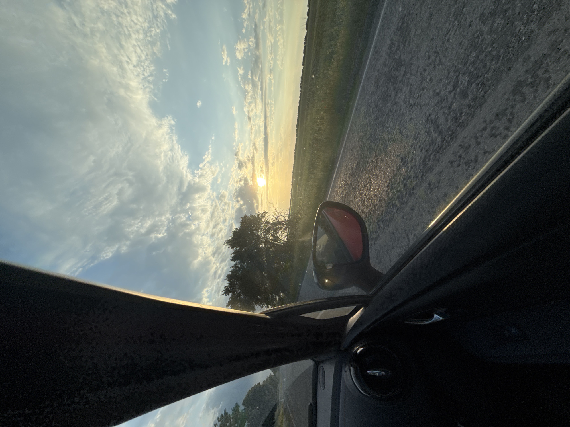 The image depicts a view from inside a car, showing a rural landscape at sunset. A road runs along the right side, bordered by a field. The sun is setting on the horizon, partially obscured by trees. A red side mirror is visible. The sky is filled with clouds. The car's interior is dark.