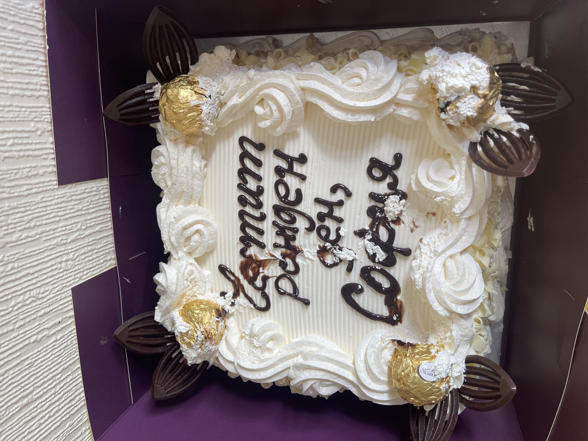 Here's a description of the image:
The image shows a cake in a purple cardboard box. The cake is square and frosted with white icing and cream swirls around the edges. There are chocolate decorations and gold-wrapped candies placed around the perimeter of the cake. In the center of the cake is writing in dark brown icing. The cake appears to be resting on a corrugated surface, possibly a wall or another part of the box.
I cannot determine the location within Maidstone, United Kingdom from the image alone.