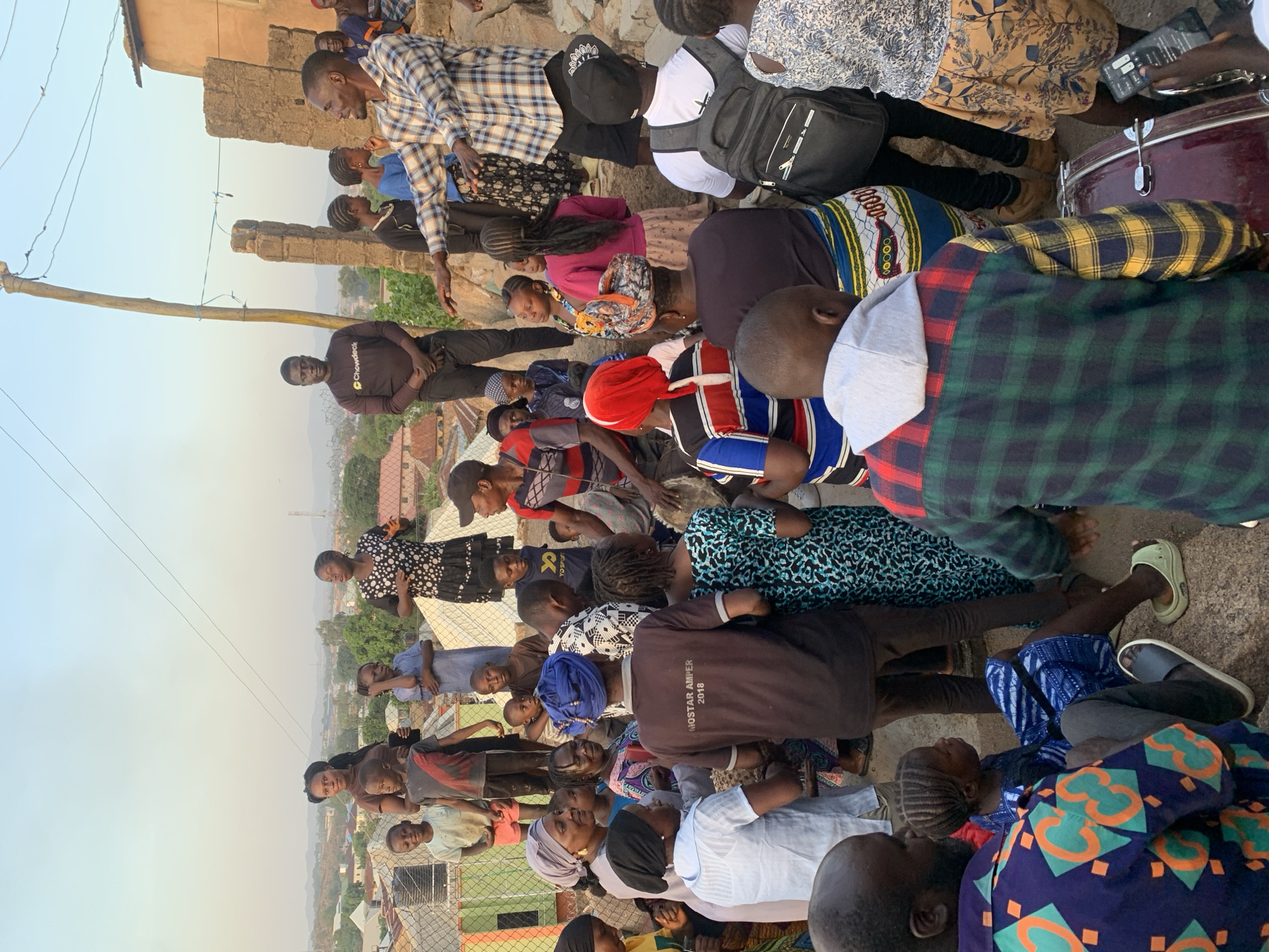 This image captures a vibrant outdoor gathering of a diverse group of people, including men, women, and children, likely in an African setting during late afternoon or early evening. The warm, soft light illuminates the scene, creating a pleasant atmosphere.
The subjects are densely packed, with individuals standing, sitting, and kneeling on what appears to be uneven, unpaved ground. Many are dressed in casual attire; notable clothing includes several plaid shirts, bright patterned fabrics, t-shirts, and head coverings. One man in a black t-shirt has the word "Chowdeck" visible on it. Another individual wears a dark hoodie with "GHOSTAR AMPER 2018" printed on the back.
The activity appears to be a communal event or meeting, with many people looking towards a central point or engaging with each other. One man in a plaid shirt is seen with his arm extended, possibly addressing the crowd. To the right, part of a red drum is visible, hinting at the presence of music or a performance.
In the background, there are various buildings, some appearing rustic or unfinished, along with sparse vegetation. A wooden utility pole with wires stretches across the left side of the frame. A chain-link fence is visible in the middle and background, separating the immediate gathering from a distant landscape featuring a valley or lower-lying residential area with more structures and hills. The overall mood appears engaged and communal.