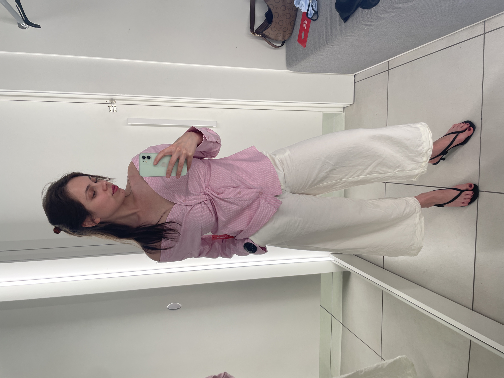 A woman is standing in a brightly lit room, likely a fitting room or a changing room, as evidenced by the mirrors and the general decor. She is taking a mirror selfie with her phone, which is light green. She is wearing a pink and white striped off-the-shoulder shirt with a knot detail in the front. Her pants are white and wide-legged. She is wearing black thong sandals with a small heel. Her hair is pulled back in a bun. In the background, there is a grey bench with a brown logo-print handbag and some clothing items on it. The floor is tiled. The location is identified as Vecsés, Hungary.