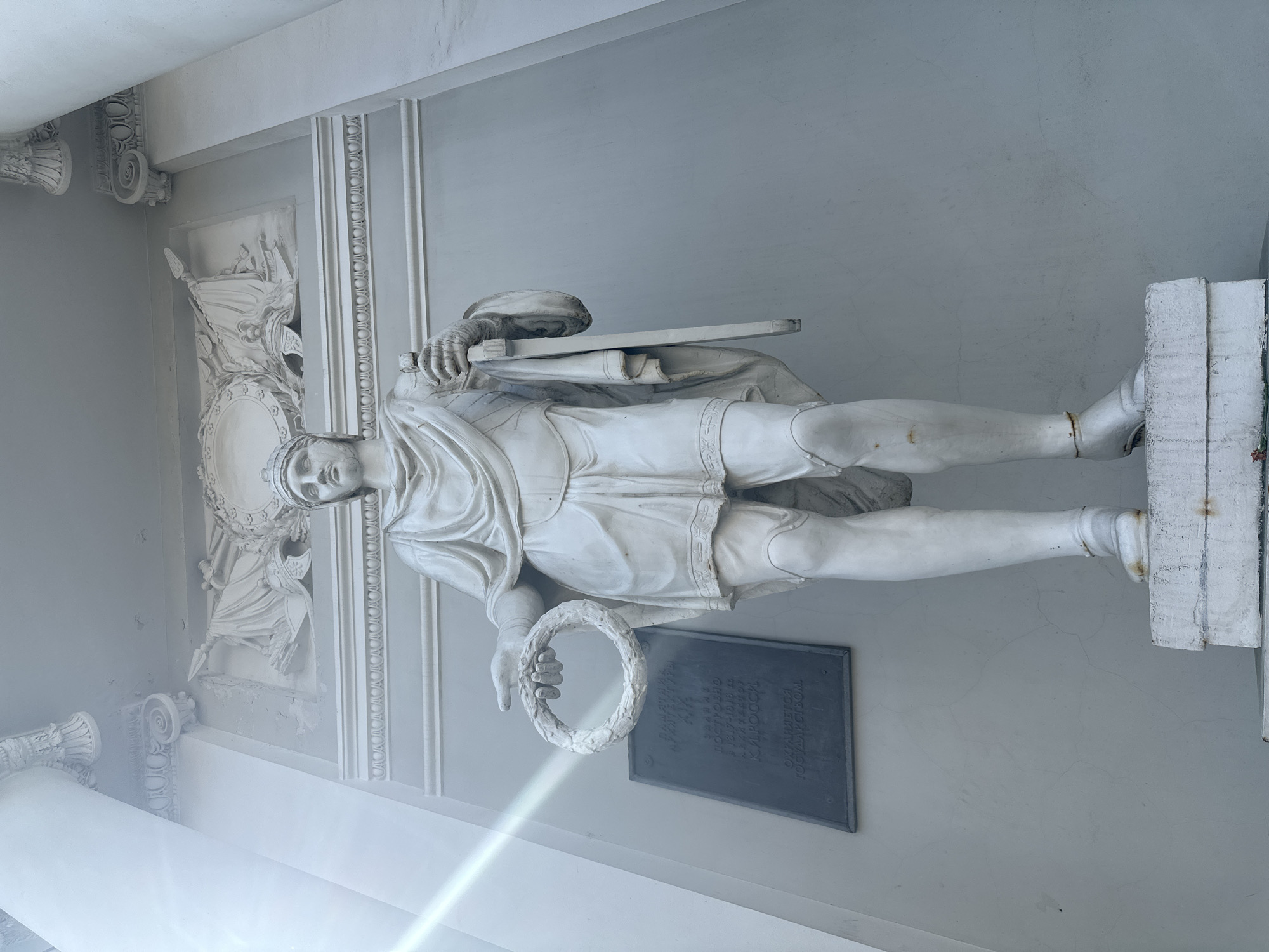 The image shows a statue of a man in a classical style, located in Saint Petersburg, Russia. The statue is white and stands next to a dark plaque. The man is wearing a toga and holding a laurel wreath in his left hand. He has a sword on his left side. The statue is set against a gray wall with decorative molding. The architecture suggests a building of historical or cultural significance.