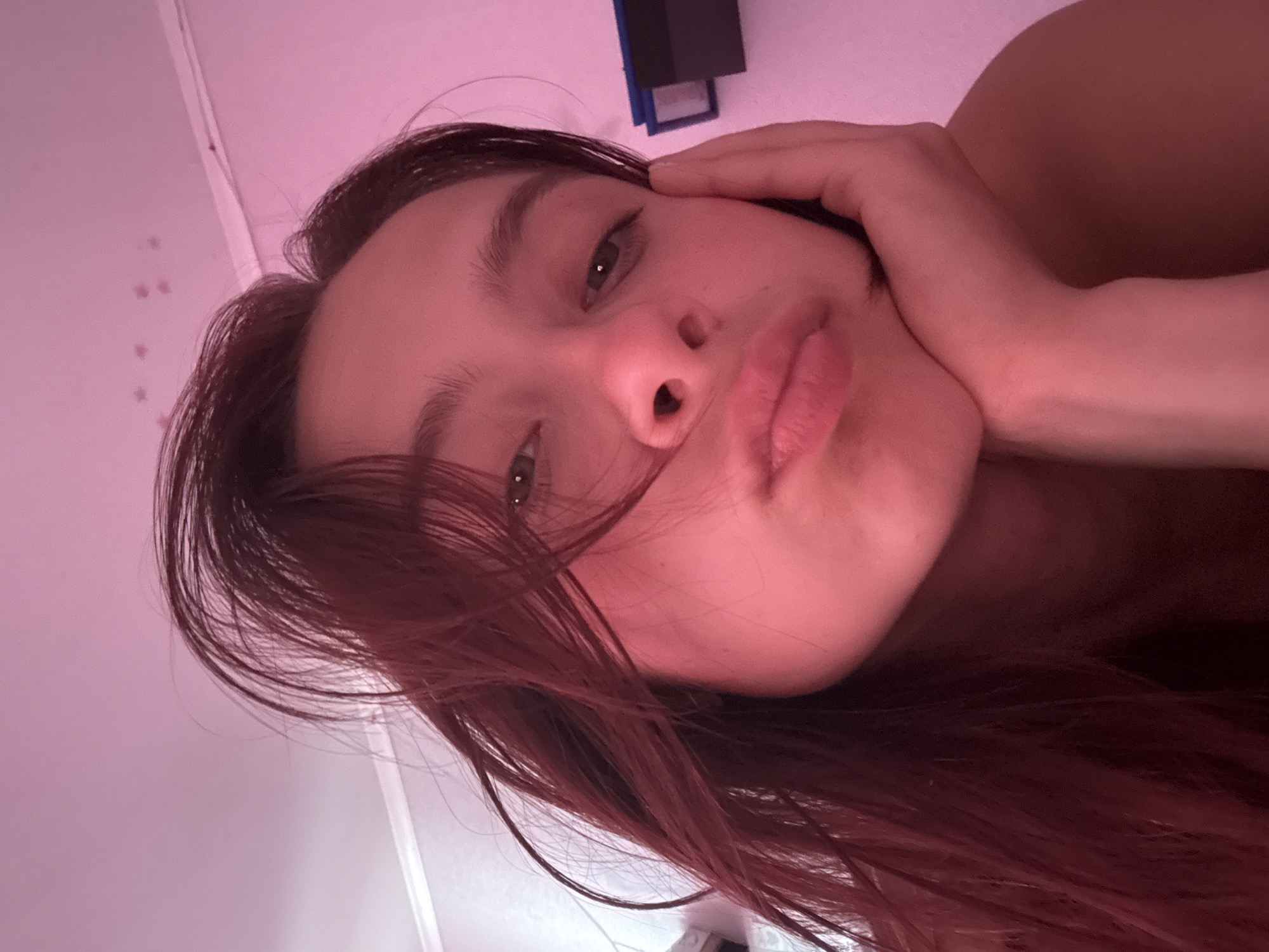 A young woman with reddish-brown hair, wearing a pink top, rests her chin on her hand. She is indoors, likely in a bedroom, with a pink-lit wall behind her. There is a dark object on the wall to the right of the frame. The image is a close-up selfie. The location provided, Прикамский, Russia, is a village in the Perm Krai region.