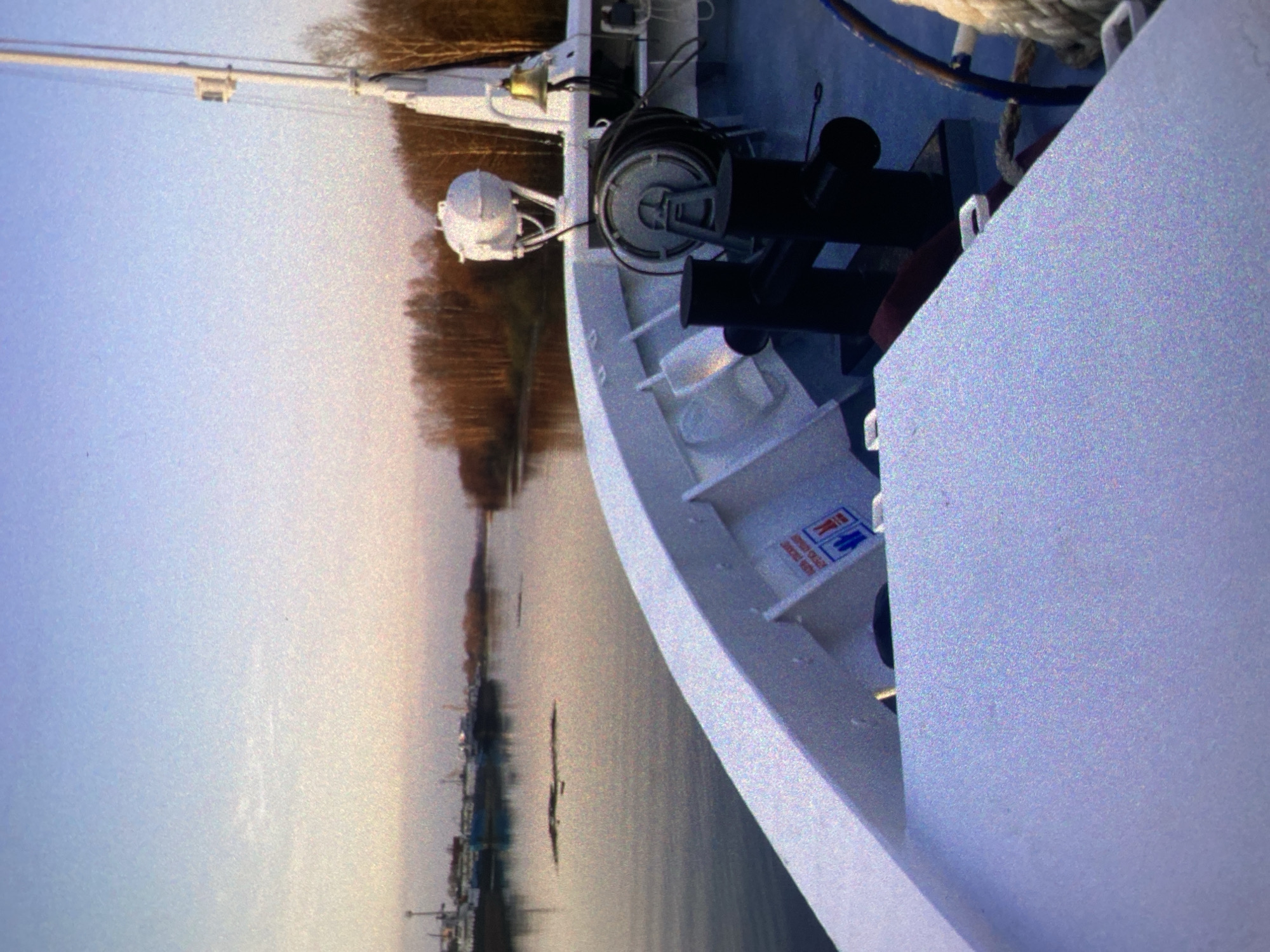 The image, oriented vertically, captures the bow of a large white vessel. The boat occupies the right half of the frame, showcasing its clean white hull and various deck fittings. These include a prominent silver-colored winch with coiled ropes, sturdy black bollards, a gleaming ship's bell, and part of a mast extending upwards.
The background, appearing to the left of the vessel due to the image's orientation, shows a calm body of water. Across the water, a distant treeline is visible, featuring bare deciduous trees, indicative of a colder season like late autumn or winter. Faint outlines of structures or other boats can be discerned along this distant bank. The sky, a hazy expanse transitioning from pale blue to a warmer, yellowish tint, occupies the far left portion of the image. No people are visible.
The vessel appears stationary on the water, likely docked or anchored. The lighting is soft and diffused, characteristic of either early morning or late afternoon, under an overcast or hazy sky. The overall image exhibits a grainy texture. Given the Saint Petersburg location context, the waterway is likely one of the city's rivers or canals.
On the white hull of the boat, near the center-right of the image, a small blue rectangular sign with a white border is visible. It features red stick-figure symbols, and below them, some white text. While largely illegible due to the image's resolution and noise, the text appears to be a short instruction, possibly related to boat operations, such as "OPEN OR CLOSE DOOR."