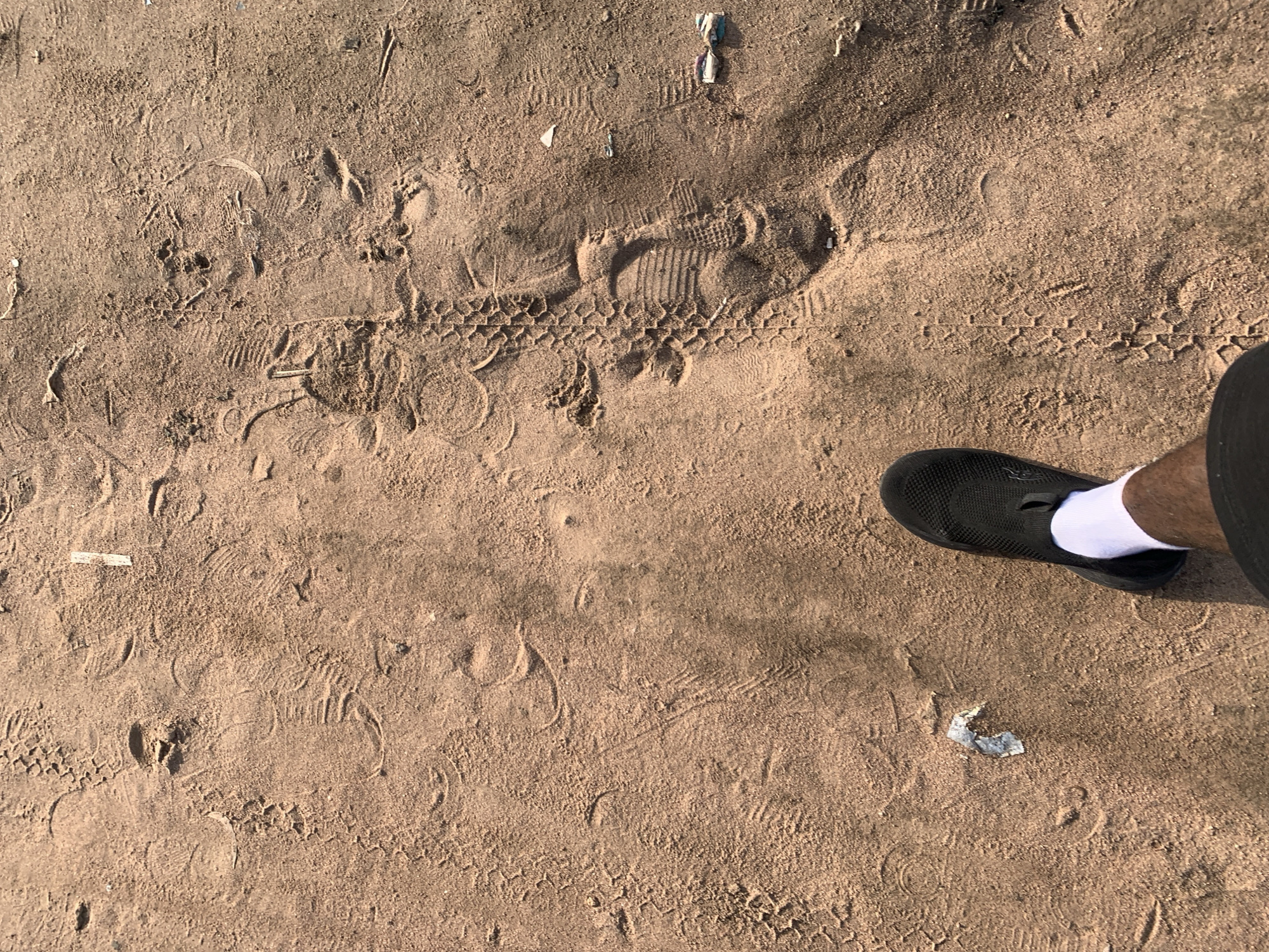 The image shows a top-down view of a sandy ground in Jalingo, Nigeria, with numerous footprints and tire tracks imprinted on its surface. In the foreground, a person's left leg is visible, wearing a black shoe and a white sock. The ground is composed of reddish-brown sand, with some small pieces of litter scattered about. The lighting suggests it is daytime, with shadows indicating the sun's position. The scene appears to be an outdoor area, possibly a path or a public space where people and vehicles have traversed, leaving behind a textured impression of their passage.