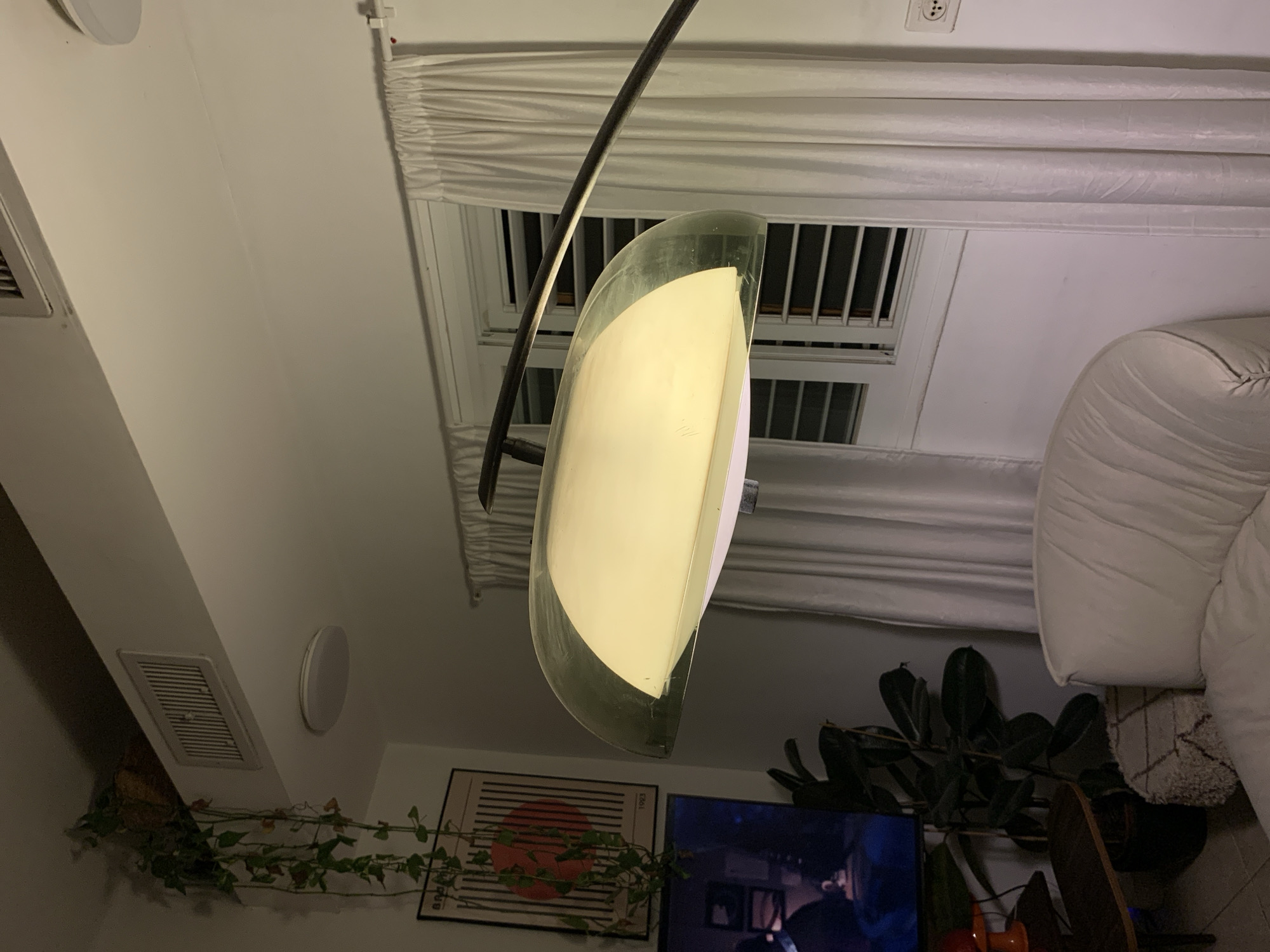 A low-angle interior shot shows a room in Tel-Aviv, Israel. A floor lamp with a translucent shade dominates the foreground. In the background, a window with white curtains, a white couch, potted plants, and a television are visible. A framed print hangs on the wall.