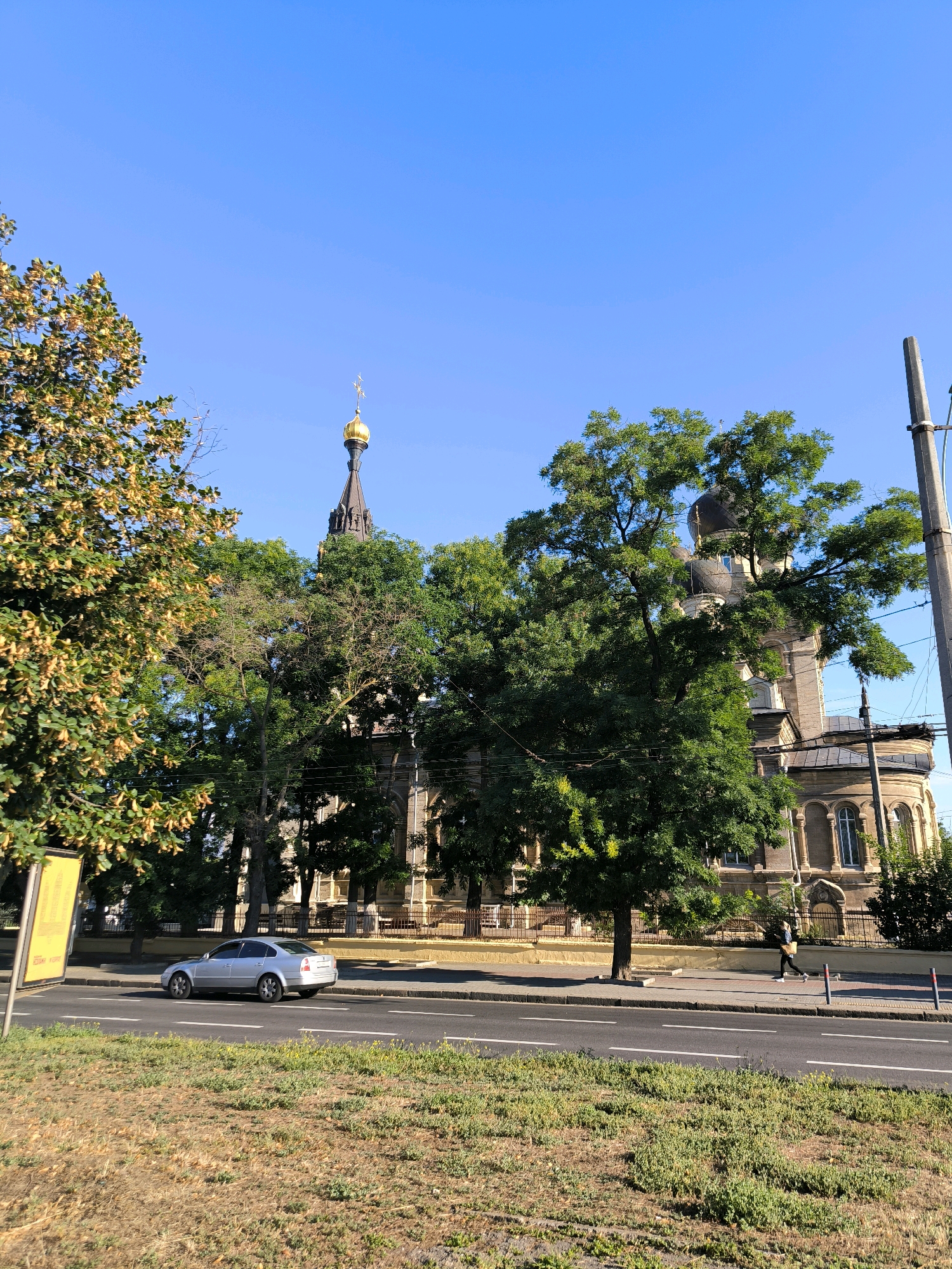 This is a street-level, eye-level, medium shot of a sunny day in Mykolaiv, Ukraine. In the background, a large stone building with domes, indicative of a church or cathedral, is partially obscured by lush green trees. A tall, slender golden dome with a cross is visible at the top of one spire. A silver sedan is driving on the road in front of the building. A pedestrian is walking on the sidewalk. The foreground shows dry, brown grass. The sky is clear and blue.