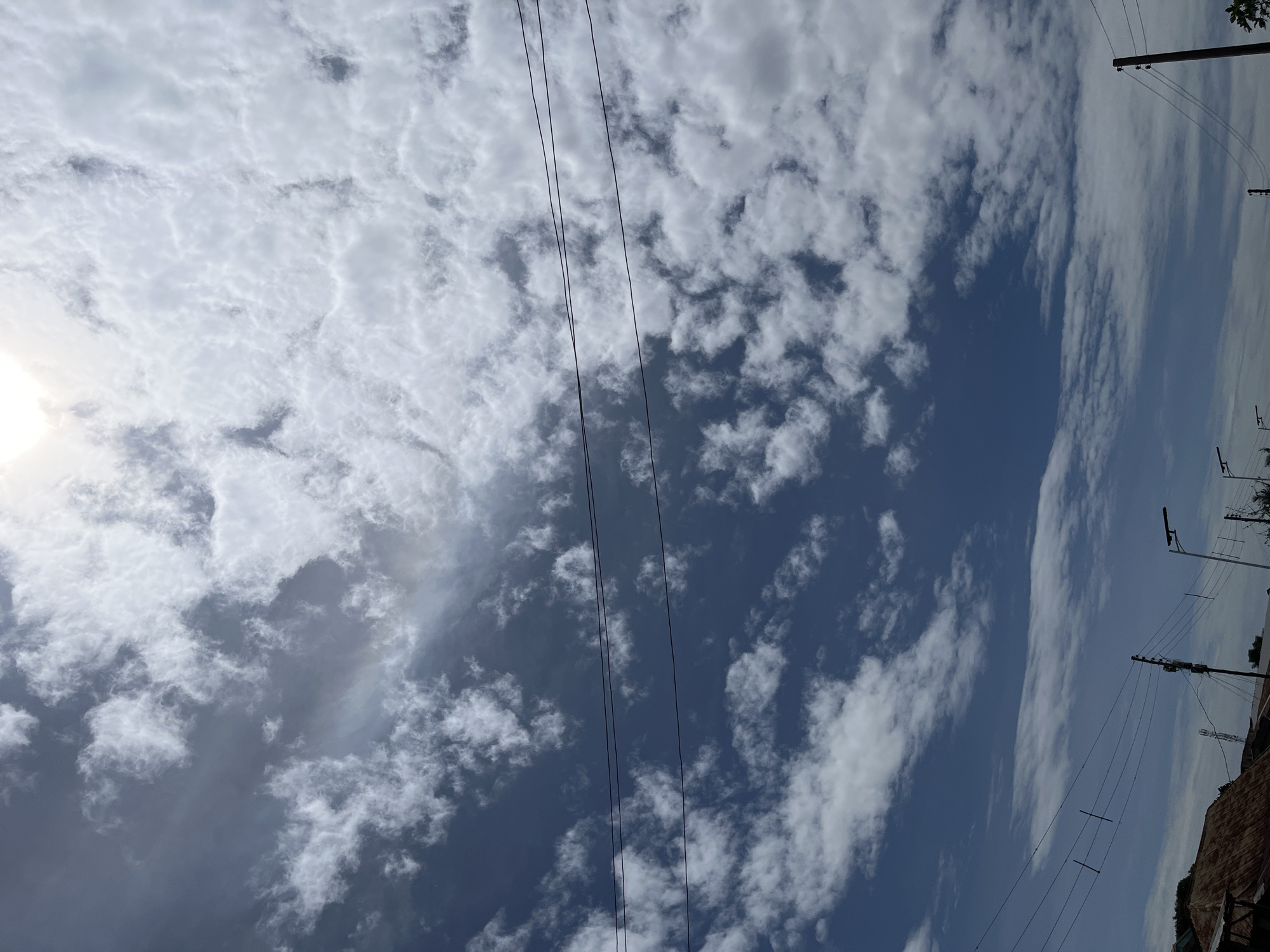 This image captures an outdoor scene in the city of Barakin Akawo, Nigeria, primarily focusing on the sky and overhead infrastructure. The upper portion of the frame is dominated by a bright, partially obscured sun, creating a significant glare in the top center. The sky exhibits a clear blue expanse interspersed with numerous white and grey clouds of varying densities, appearing as cumulus and stratocumulus formations. A subtle iridescent or halo-like effect is faintly visible within the clouds in the upper-mid left quadrant.
Horizontally spanning the middle and lower sections of the frame are multiple dark, thin utility lines, likely power or communication cables. These lines are supported by several dark, vertical utility poles, visible mostly in the lower half of the image. Some poles feature attached streetlights. In the bottom left corner, a portion of a building with a corrugated, rusted metal roof is visible. Further along the lower edge, indistinct building structures and sparse vegetation are partially discernible. No individuals or specific activities are present in the frame.