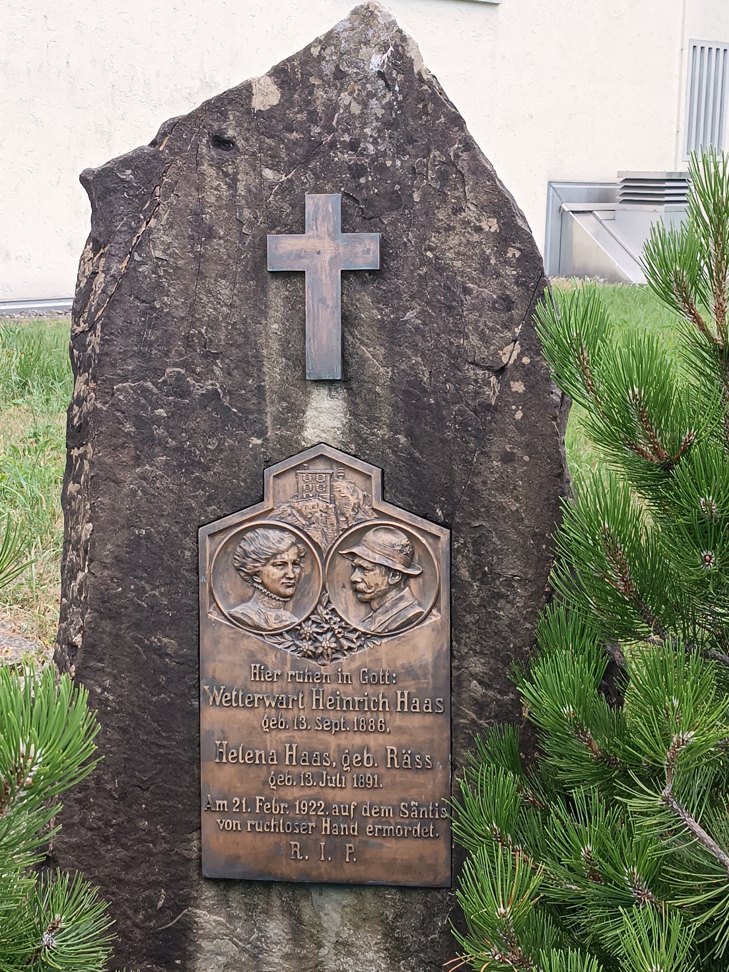 The image shows a tombstone in Appenzell, Switzerland, with a bronze plaque featuring portraits of a man and a woman. The plaque reads: "Hier ruhen in Gott: Wetterwart Heinrich Haas, geb. 13. Sept. 1886. Helena Haas, geb. Räss, geb. 13. Juli 1891. Am 21. Febr. 1922, auf dem Säntis von ruchloser Hand ermordet. R. I. P." This translates to: "Here rest in God: Weather observer Heinrich Haas, born Sept. 13, 1886. Helena Haas, née Räss, born July 13, 1891. On Feb. 21, 1922, murdered on the Säntis by a ruthless hand. Rest in Peace." The tombstone is a large, rough-hewn stone with a metal cross affixed to its upper portion. Pine branches frame the right side of the image, and green grass surrounds the base of the tombstone.