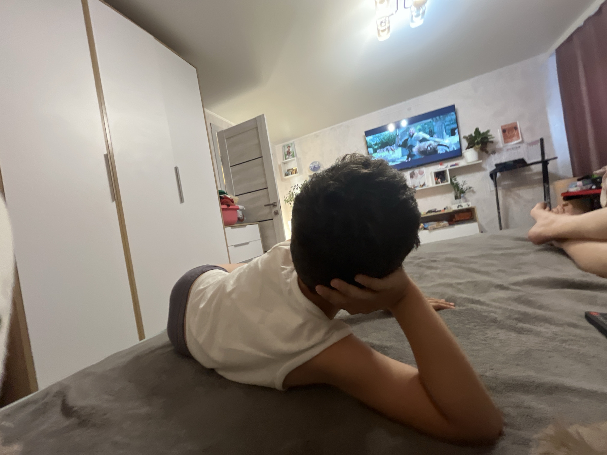 A young boy is lying on his stomach on a bed, facing away from the camera and looking towards a television that is playing a movie. He is wearing a white t-shirt and dark grey shorts. His right arm is bent and supporting his head, and his left arm is extended on the bed. In the background, there is a large white wardrobe, a door, a television on the wall, shelves with decorations, and a purple curtain. The scene appears to be a bedroom in a residential building. The location is specified as Krasnyye Barrikady, Russia.