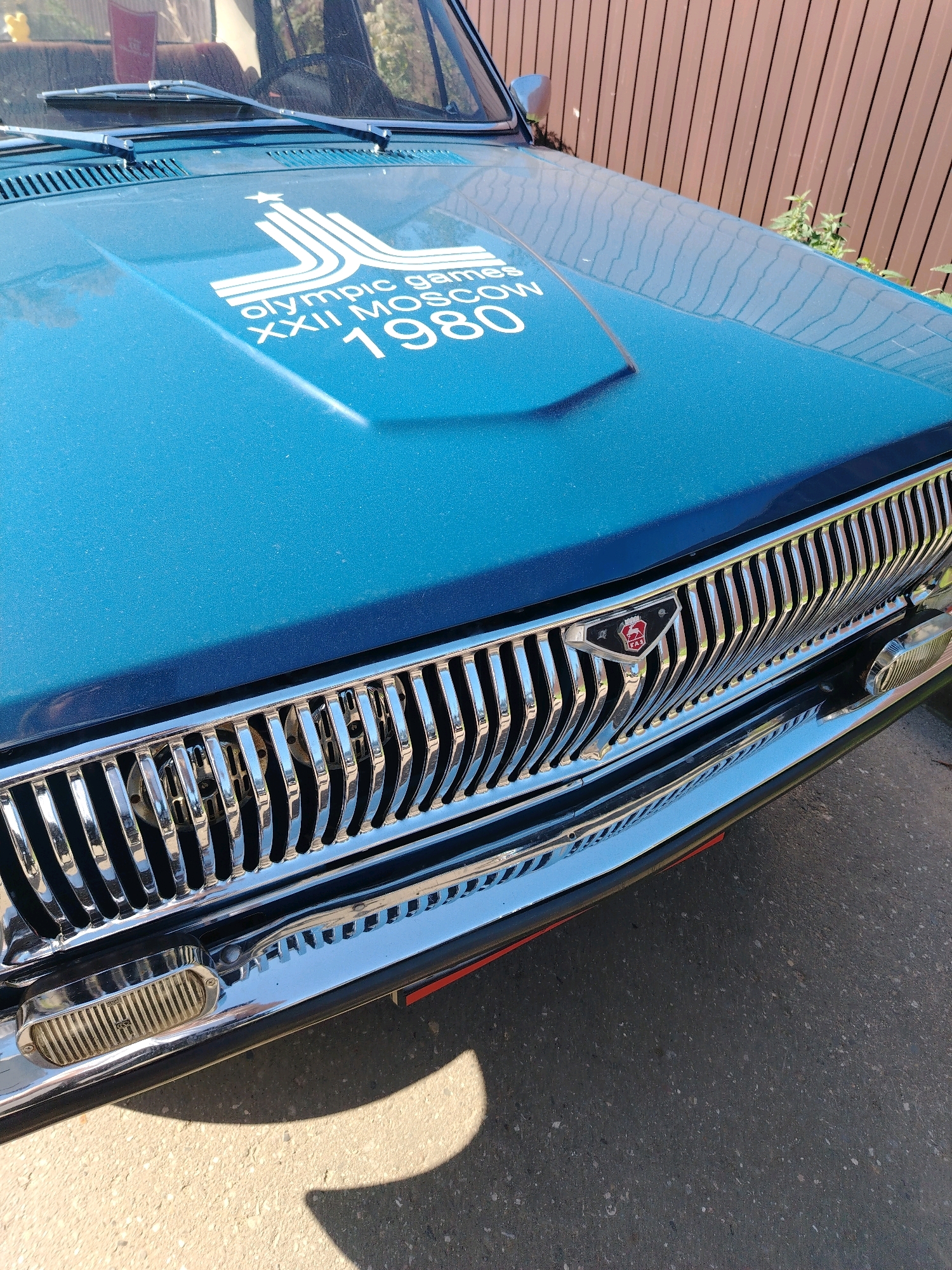 The image shows the front of a teal-colored car with a chrome grille. On the hood of the car, there is a white graphic that reads "OLYMPIC GAMES XXII MOSCOW 1980". The car is parked on an asphalt surface, and a wooden fence is visible in the background. The lighting suggests it is daytime. There are no people visible in the image. The image does not contain any information that would indicate its location in Yaroslavl, Russia.