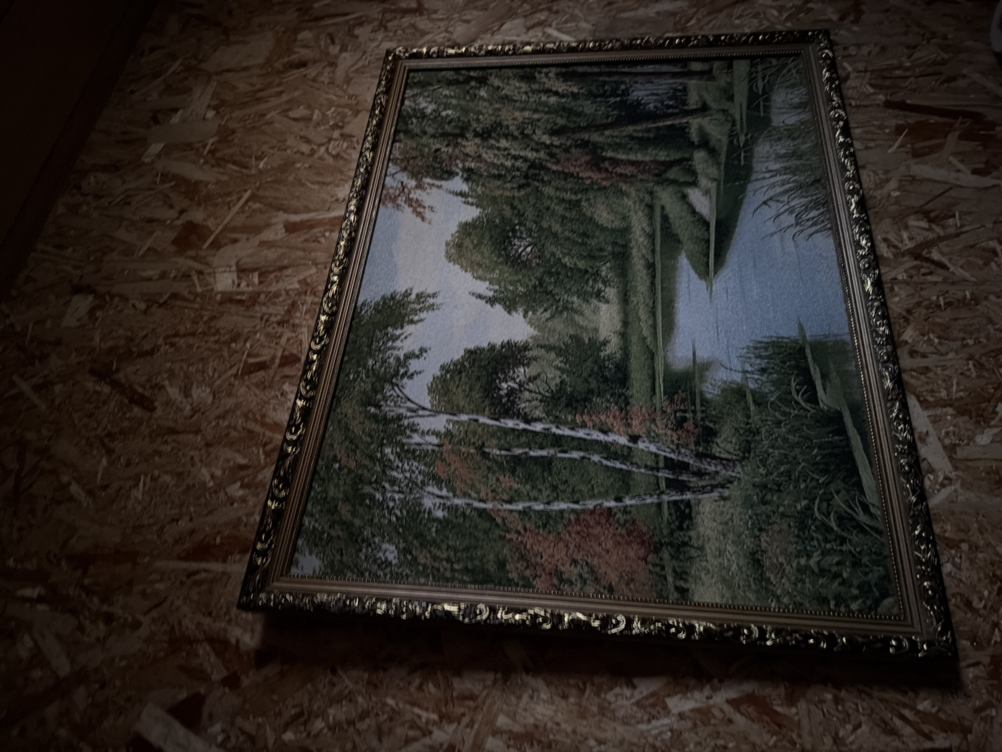 A framed landscape painting hangs on a wall constructed of oriented strand board (OSB). The painting depicts a tranquil scene with trees, water, and foliage in various shades of green, brown, and red. The frame is ornate and appears to be made of metal. The painting is hung at an angle, and the lighting is dim, suggesting the photo was taken indoors. The location is not discernible from the provided visual information. Therefore, determining whether the location is in the city of Кадряково, Russia, is impossible.