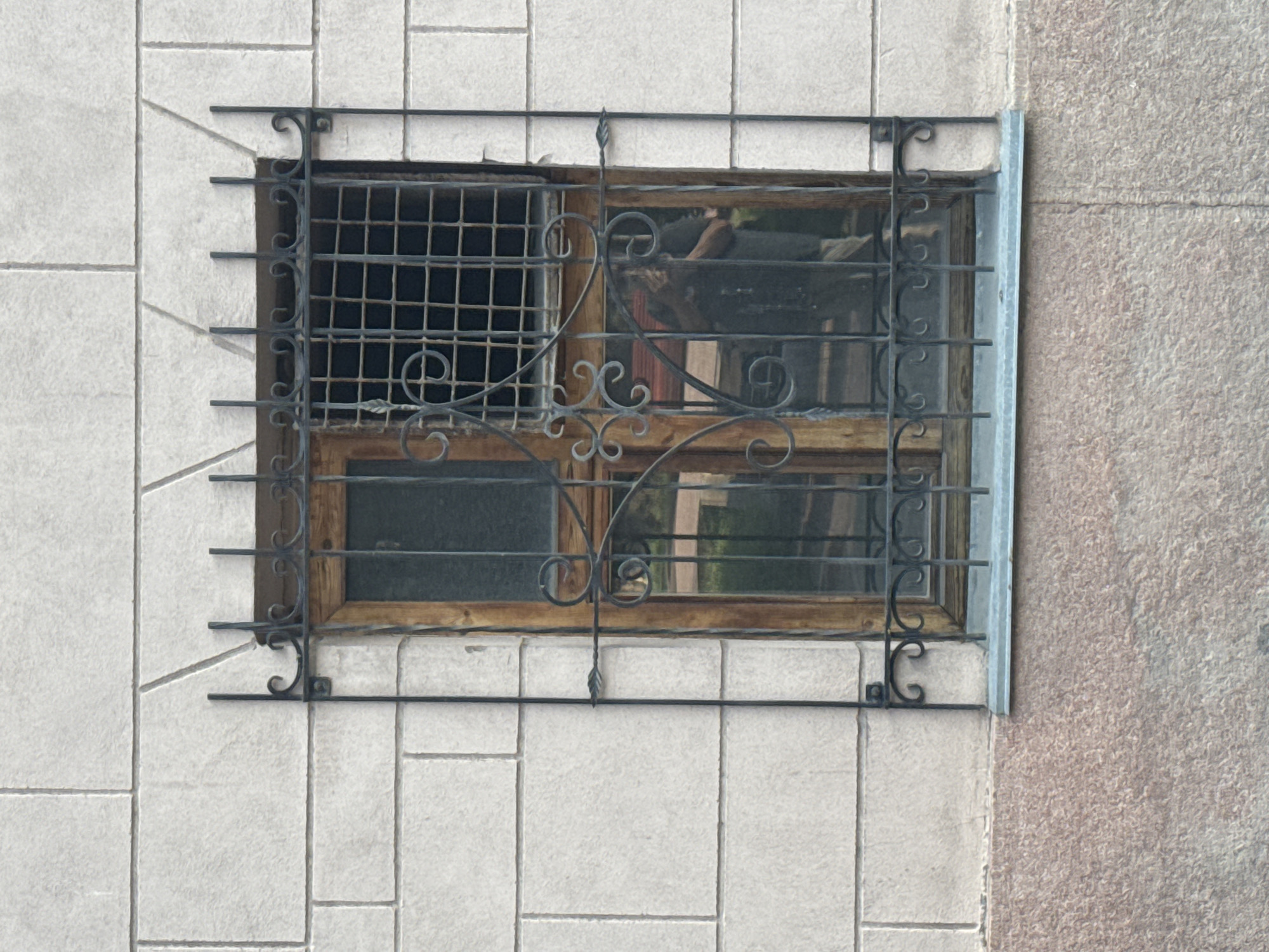 A window with ornate black metal bars is set into a light grey, block-textured building wall. The window frame appears to be made of weathered wood. The glass reflects an interior scene that includes a person, partially obscured by the bars. The building has a rough, pinkish-brown lower section. This image was taken in Bishkek City, Kyrgyzstan.