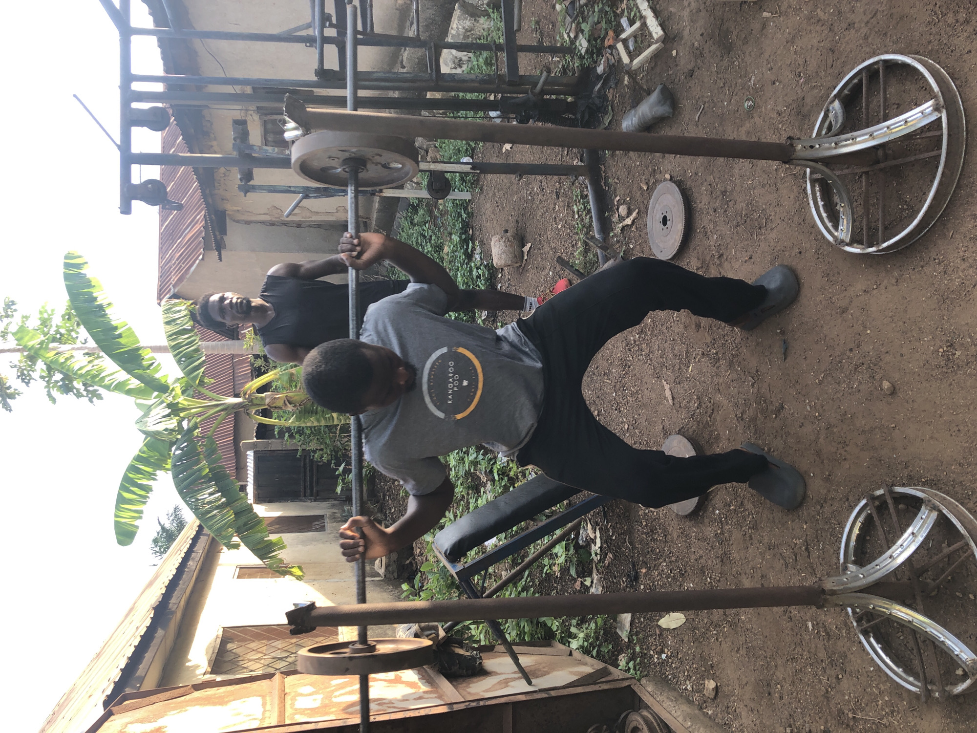 The image depicts two men in an outdoor setting, likely Akwanga, Nigeria. One man is actively engaged in weightlifting using a makeshift bench press constructed from metal components and repurposed items. The weights appear to be composed of circular metal plates. The man is wearing a grey t-shirt and dark pants.
Another man stands behind the weightlifter, observing and smiling. He is wearing a dark-colored sleeveless shirt.
The environment includes various metal structures, a bench, and vegetation. The ground is dirt. The presence of a banana tree suggests a tropical climate consistent with the location. The background features buildings with corrugated metal roofing.