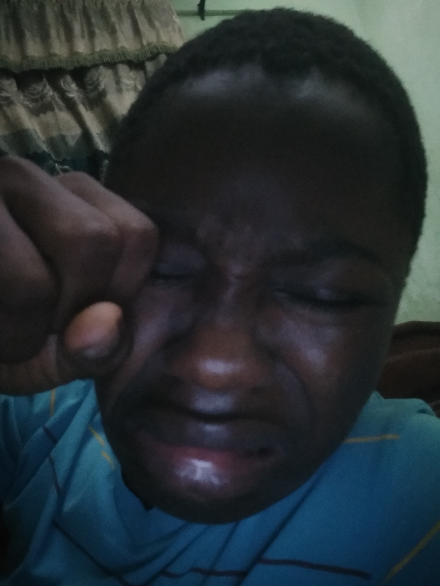 A dark-skinned person is clenching their fist near their face with their eyes closed and mouth contorted. They are wearing a blue shirt with stripes. A curtain is visible in the background. The location is unknown.