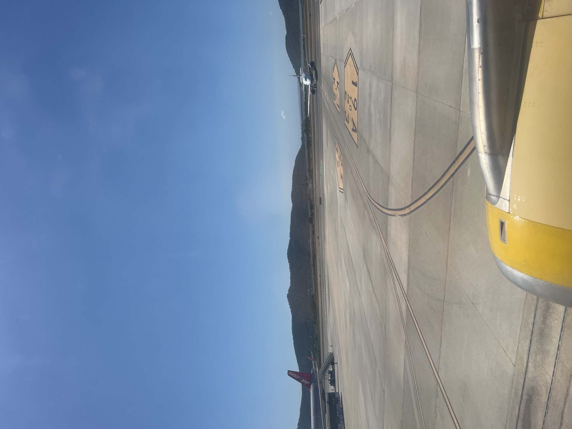 The image shows an airport tarmac on a clear, sunny day. In the foreground, a portion of a yellow and silver airplane engine is visible. The tarmac is marked with yellow lines and numbers, indicating taxiways and runway designations. Two airplanes are present on the tarmac. On the left, a red and white airplane with "jet2.com" branding is parked with stairs attached. Further in the distance, on the right, a white airplane is on the tarmac. Rolling hills covered in green vegetation form the background, with a clear blue sky overhead. This scene is consistent with an airport located in or near Sant Josep de sa Talaia, Spain, given the climate and geographical features. There are no people visible in the image.