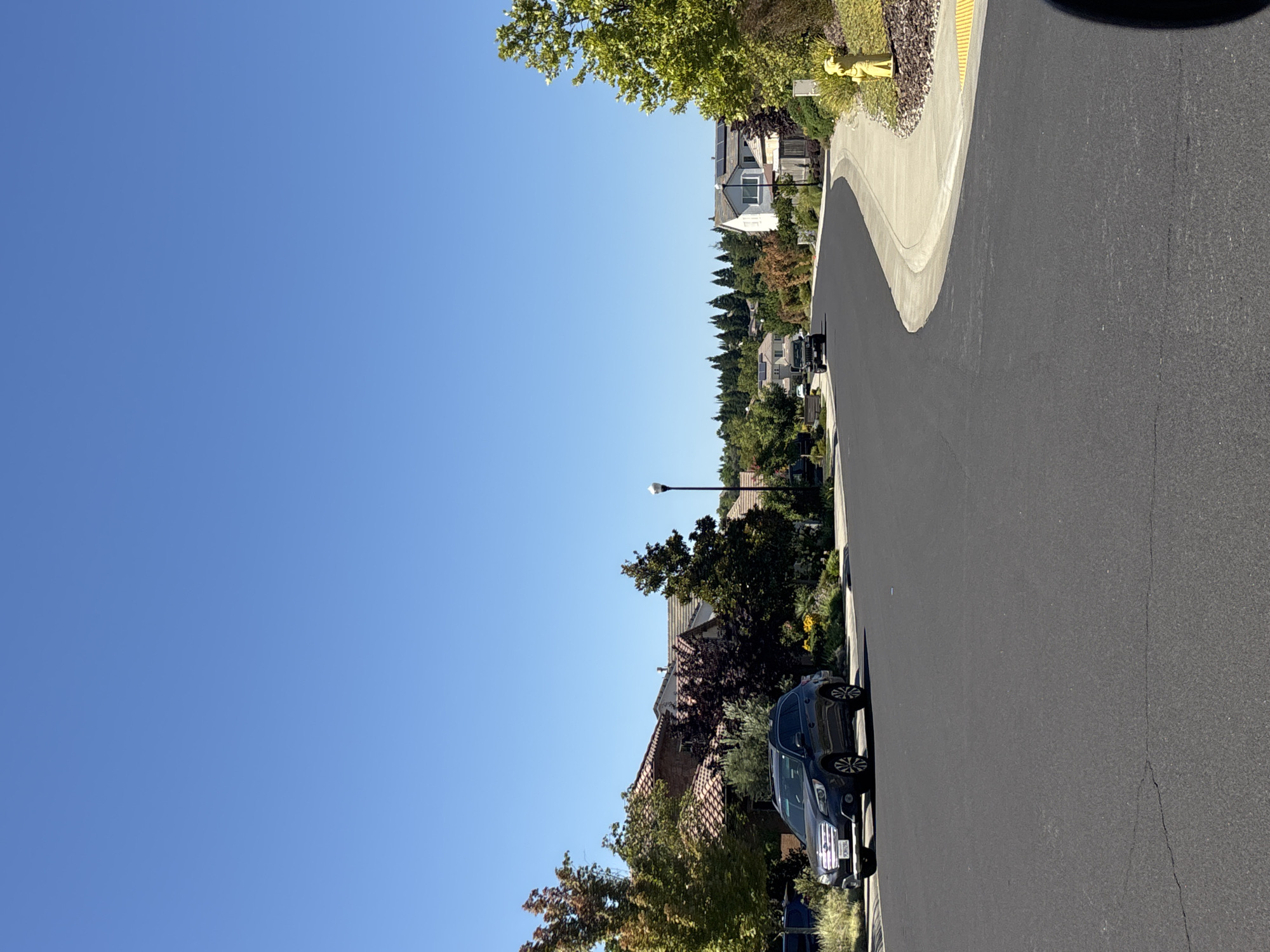 This is an exterior, eye-level, medium shot of a suburban street in Roseville, California, on a clear, sunny day. The foreground is dominated by a newly paved asphalt road curving to the right. A dark blue Subaru Outback is parked on the left side of the road. The background features a row of houses with tiled roofs and well-maintained landscaping, interspersed with trees. A street lamp is visible on the left side of the road, and a yellow fire hydrant stands on the right side of the sidewalk. In the distance, more houses and trees are visible, with a gently rolling, tree-covered hill forming the horizon. The sky is a clear, bright blue, with no clouds.
