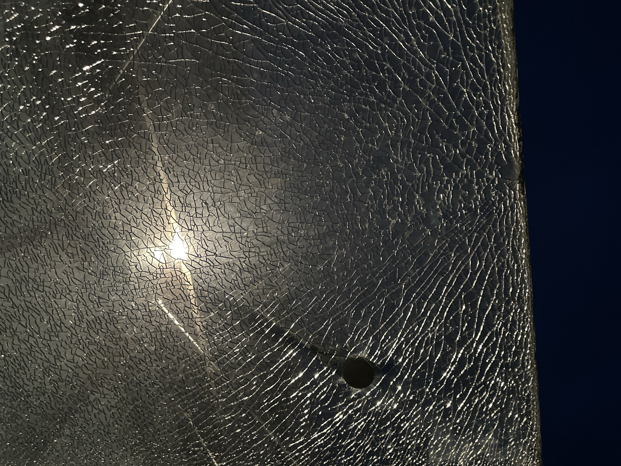 A panel of shattered glass is seen against a dark blue background. A bright light shines through the center of the glass, creating a starburst effect. A hole is visible in the lower right portion of the glass. The location is Unknown, Unknown.