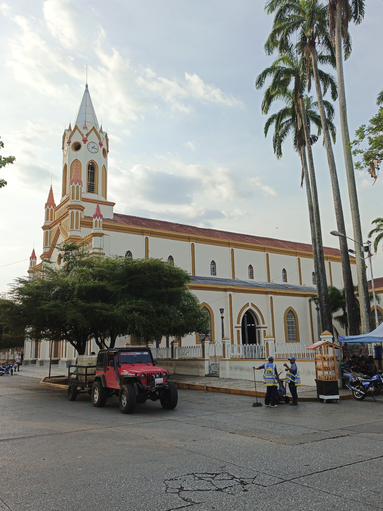 The image shows the Iglesia Inmaculada Concepción in Tinaquillo, Venezuela. A red Jeep with a trailer is parked on the street in front of the church. Two sanitation workers in blue vests are standing and conversing beside a snack cart, with a broom leaning against one of them. A motorcycle is parked near the snack cart, and people are seated under a blue tent behind it. Tall palm trees are visible to the right of the church. The sky is partly cloudy. The church is predominantly white with gold trim, a red roof, and a tall, ornate steeple with clocks on each face. A large, full tree partially obscures the view of the church's facade.