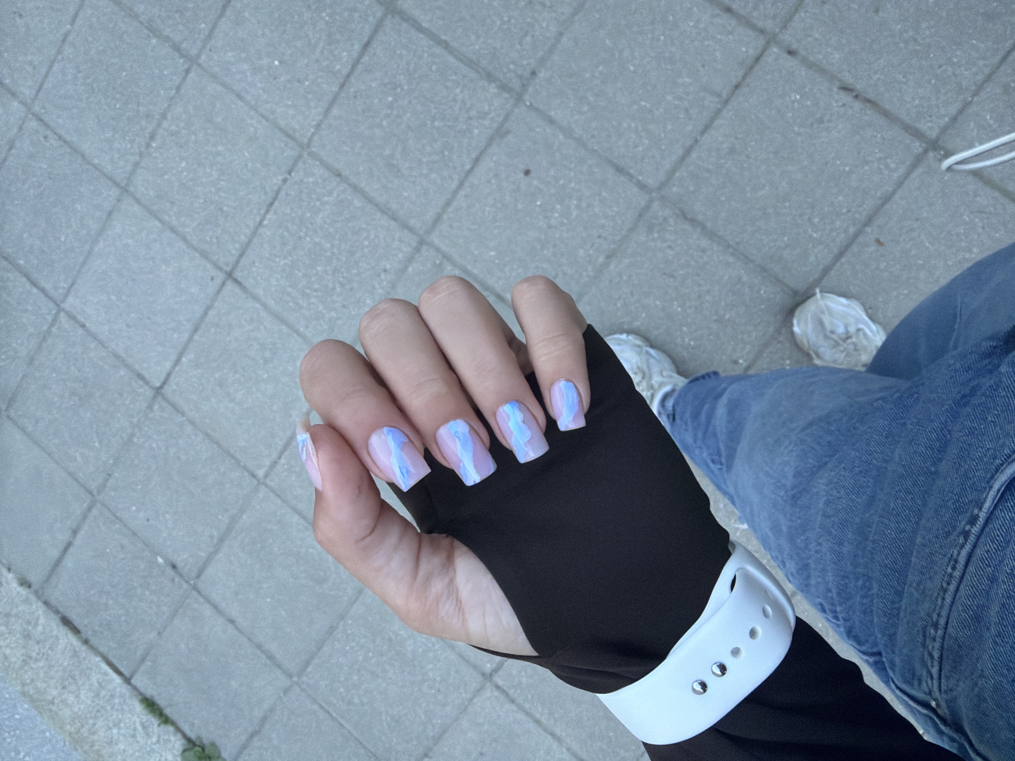 A woman's hand with pale skin and manicured nails featuring a light pink base with a blue and white marble pattern is displayed against a backdrop of gray concrete pavers. The woman is wearing a black long-sleeved shirt with a cut-out for the thumb and a white smartwatch with a silicone band. Part of her blue jeans and white sneakers are visible in the lower right of the frame. The setting appears to be an outdoor pedestrian area. It is impossible to determine if this image was taken in Gabrovo, Bulgaria without additional information.