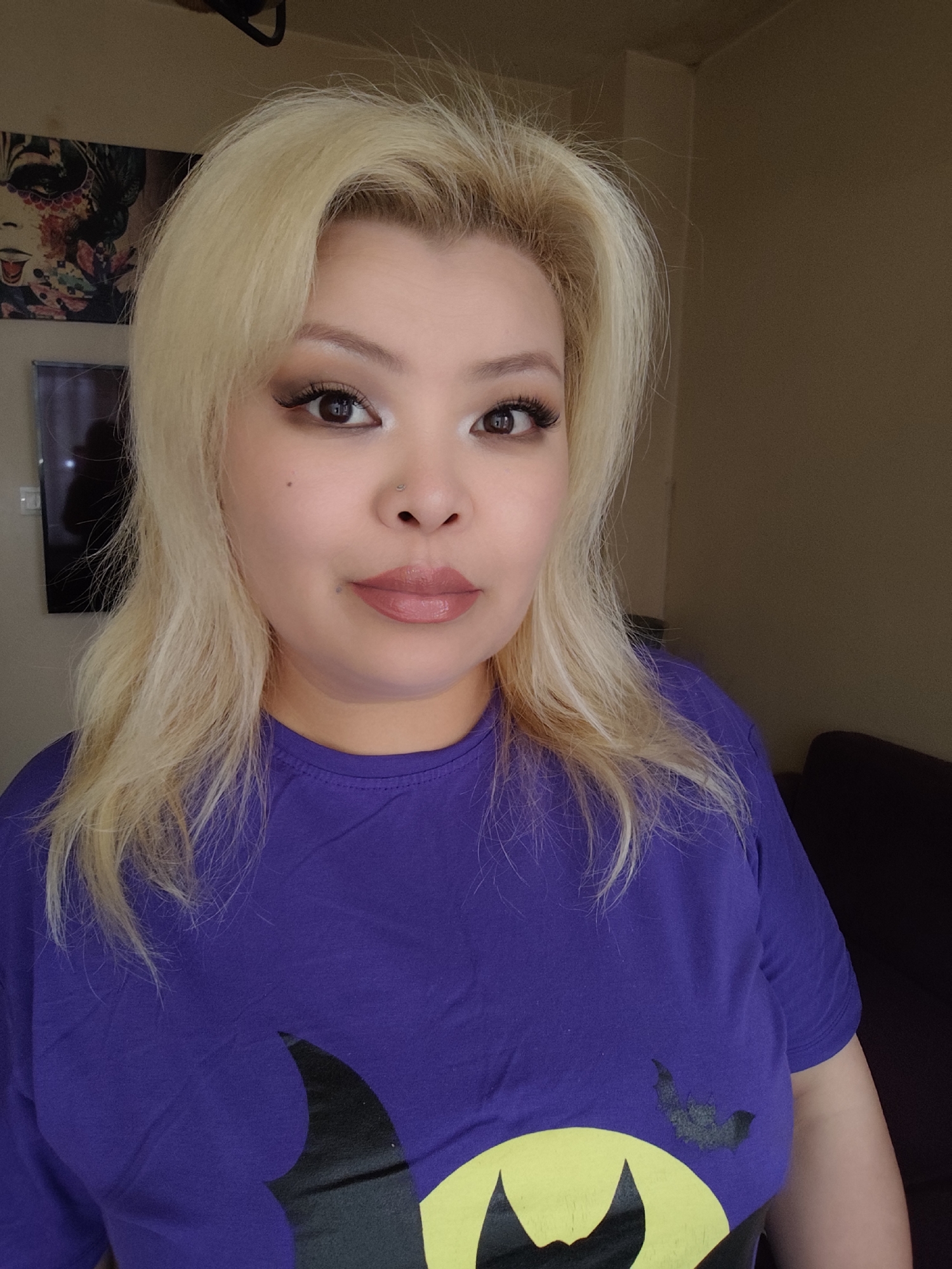 A medium shot depicts a woman with light blonde hair, wearing a purple t-shirt with a stylized Batman logo, facing the viewer. The logo is predominantly yellow with black bat wings. The woman has a light complexion, brown eyes, and a piercing in her left nostril. She is wearing makeup, including eyeshadow, eyeliner, and lip color. Behind her, out of focus, are details of a room, including a piece of artwork on the wall and a television. The location, Sarıyer, Turkey, cannot be inferred from the image.
