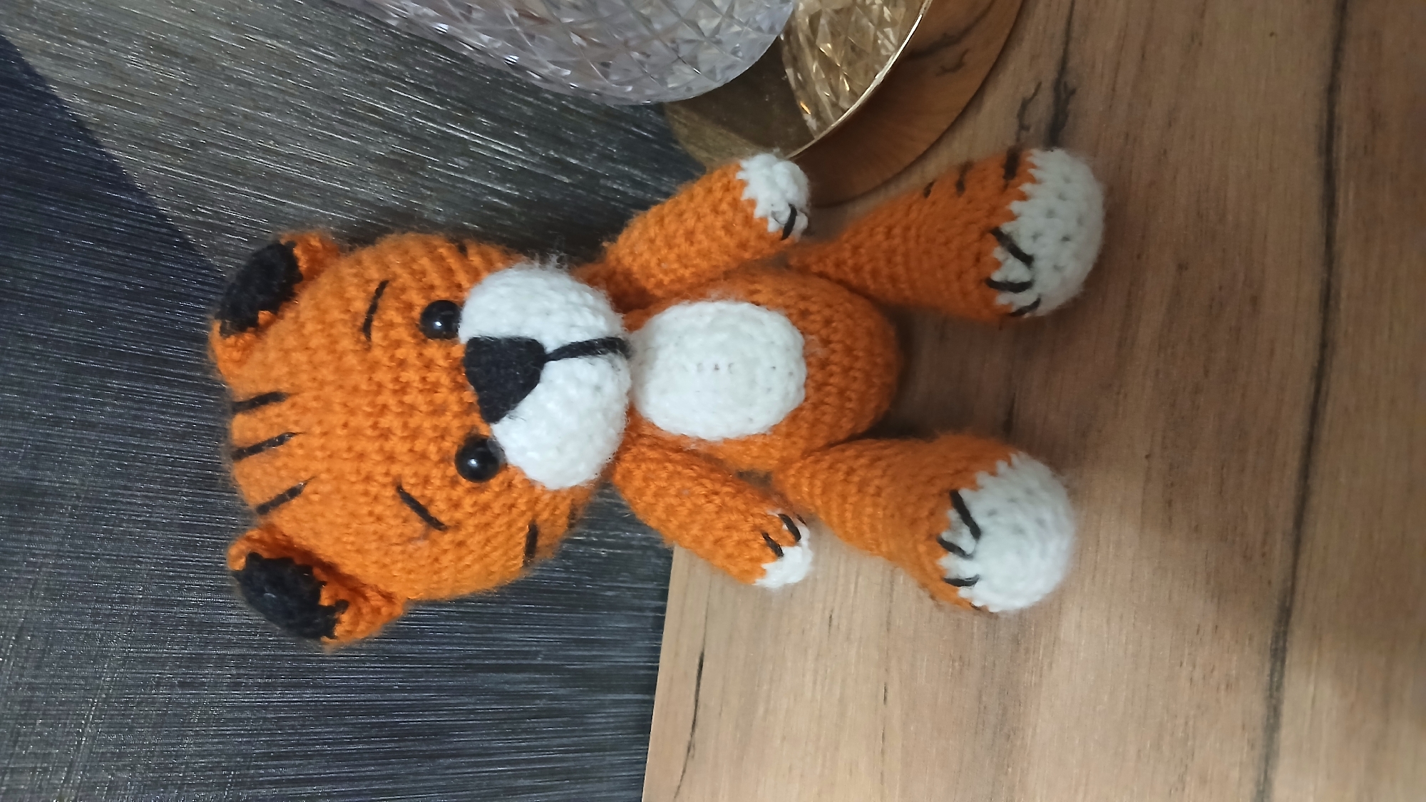 A crocheted tiger toy is positioned centrally on a wooden surface, facing forward. The tiger is orange with black stripes on its head and ears, and a white chest and muzzle. It has black button eyes and a black nose. Its paws have white cuffs with black markings resembling claws. To the right of the tiger, partially visible, is a gold-colored object with a textured, clear glass element. The background features a dark, textured surface. The scene is indoors, on what appears to be a table or shelf. The lighting suggests an indoor setting, possibly a home environment.