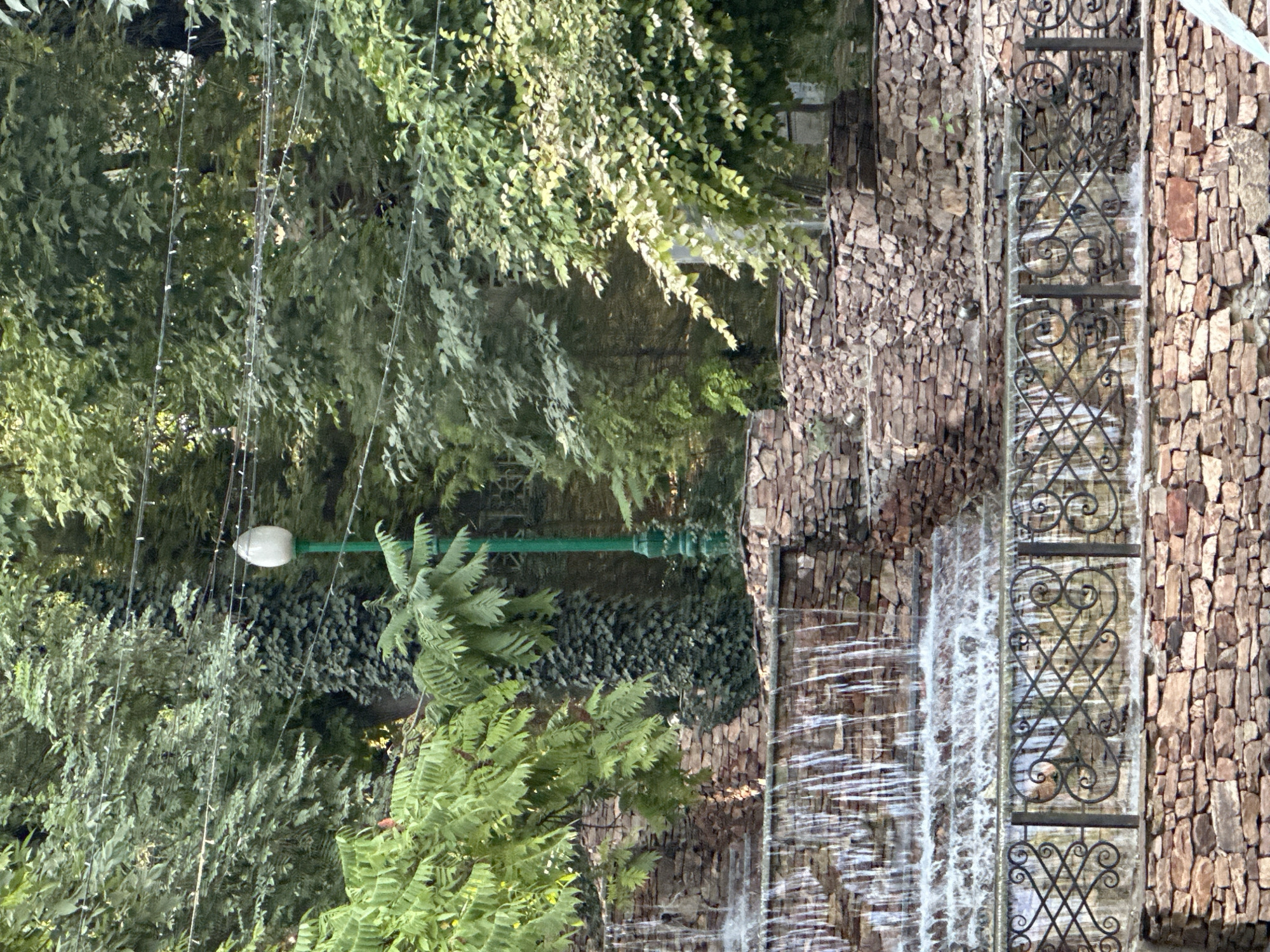 The image depicts a terraced waterfall feature constructed with natural stone in Tashkent, Uzbekistan. The waterfall cascades down multiple levels, with water flowing over rough-hewn rocks. A black wrought-iron fence with an ornate scrolling pattern separates the lower section of the waterfall from a stone wall. Lush green trees and foliage surround the waterfall, creating a natural, serene environment. A vintage-style lamppost with a white, egg-shaped lantern is visible amidst the greenery. The scene appears to be a public park or garden, likely a well-maintained leisure area within the city. The time of day appears to be daytime, with natural light illuminating the scene. There are no people visible in the image.