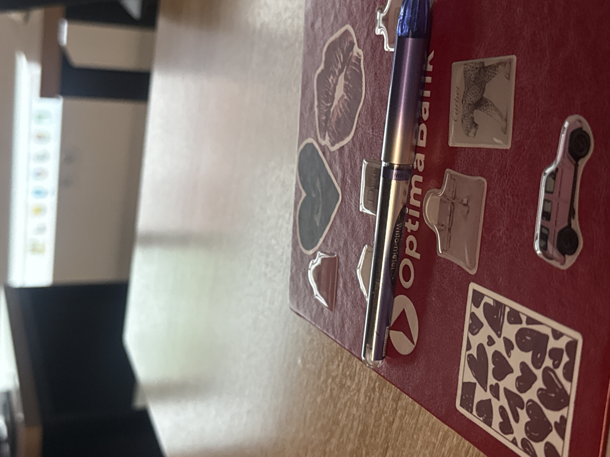 The image shows a close-up view of a red notebook lying on a wooden table. The notebook is decorated with several stickers, including a heart, lips, a handbag, a leopard print, a car, and a pattern of hearts. A purple pen with a silver tip rests on top of the notebook. The background is blurred, showing a desk and a hint of what appears to be office equipment. There are no people visible in the image. The location in Bishkek City, Kyrgyzstan is not determinable from the visual information provided.