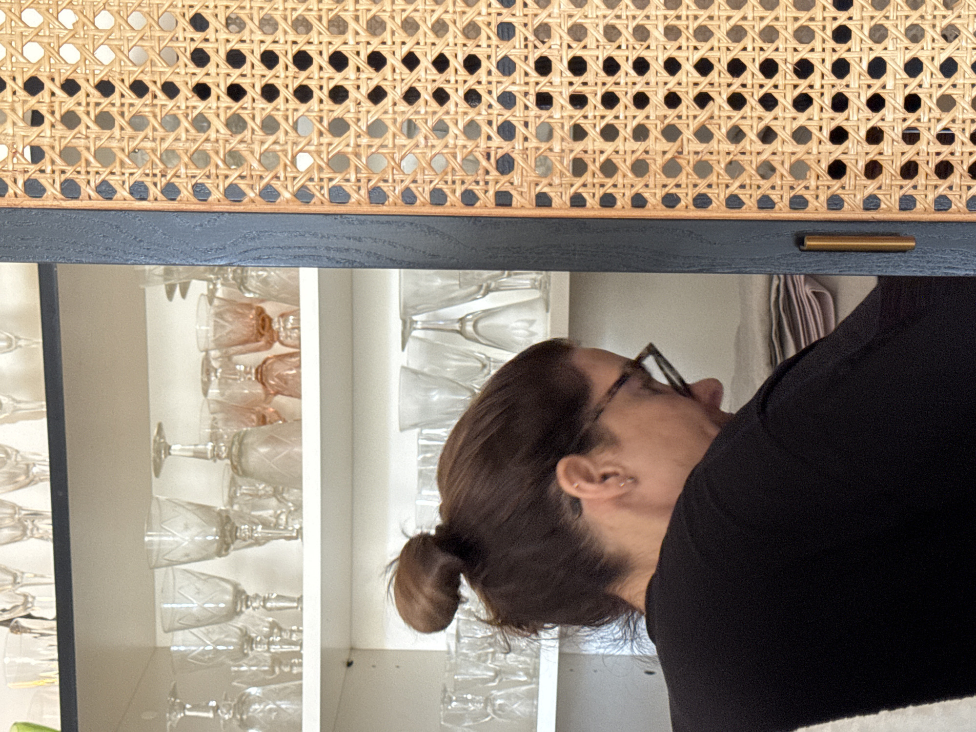 A photograph taken indoors shows a woman, viewed from the side, bending towards a cabinet. The cabinet has white shelves filled with clear and pink glassware. A drawer sits above, featuring a woven cane front, dark blue paint, and a gold pull. The woman has brown hair styled in a low bun and is wearing glasses and a black shirt. The location is unidentifiable.