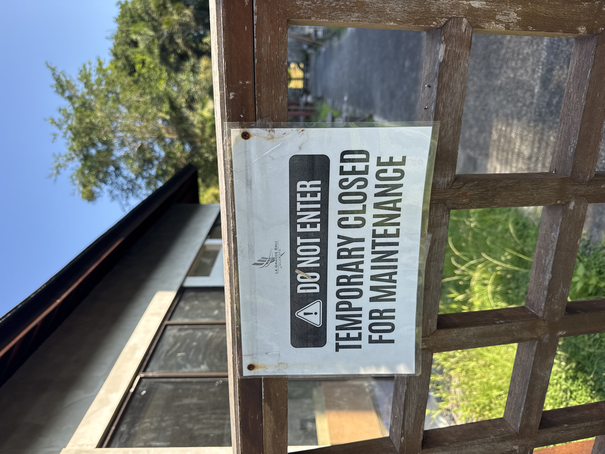 A close-up, medium shot depicts a wooden lattice fence with a sign reading "DO NOT ENTER TEMPORARY CLOSED FOR MAINTENANCE" affixed to it. The sign also includes the name "LE GRANDE BALI ULUWATU," indicating the location is in Kuta Selatan, Indonesia. In the background, a building with large windows and a sloping roofline is partially visible. A tree and a clear blue sky are visible in the upper left. The lighting suggests it is daytime, with clear visibility. The fence appears aged with weathered wood.