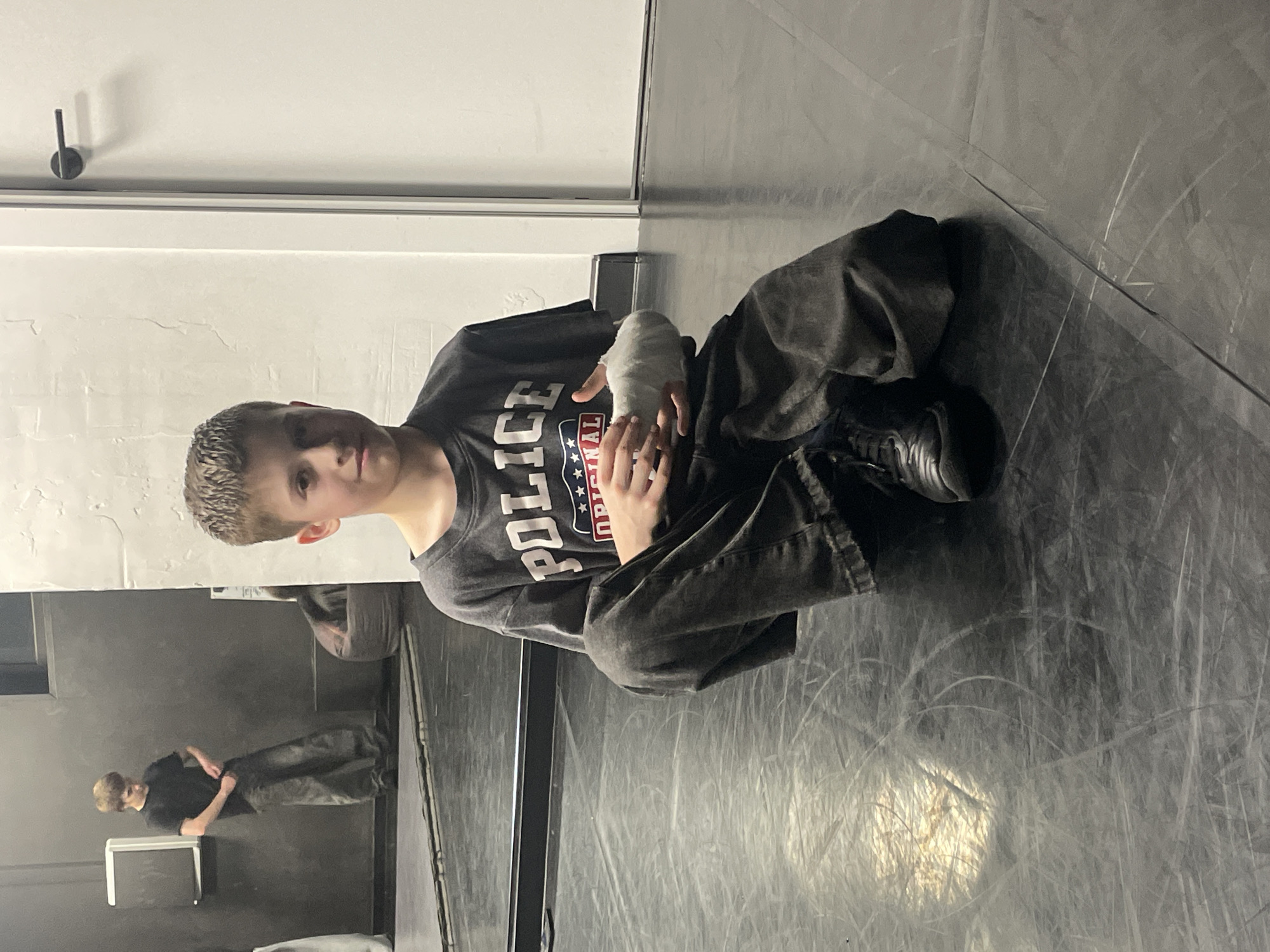A young person with short blond hair is seated on the floor with one hand wrapped in a white bandage. The person wears a dark t-shirt with the word "POLICE" and a graphic design on the front, and dark-colored pants and shoes. A mirror reflects the person and the surrounding environment. The location appears to be a room with a white wall, a dark floor, and a metal fixture on the wall. This does not provide information to identify the location as Krasnoyarsk, Russia.