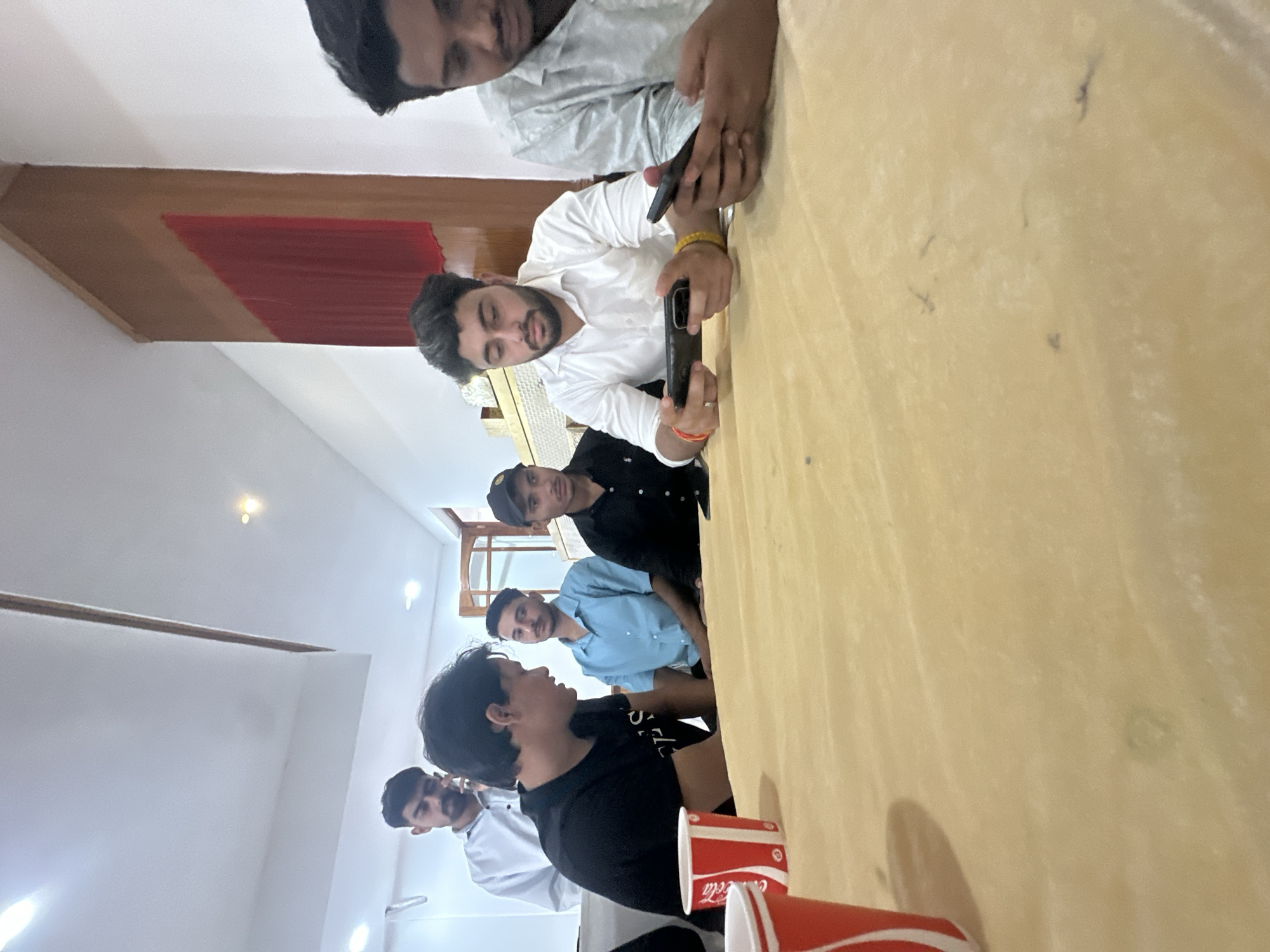 Six young men are gathered around a light brown table in a room with white walls and a light-colored ceiling. The room appears to be indoors, possibly a dining area or conference room. A tall wooden panel with a red fabric insert is visible on the right wall.
From left to right, the individuals are:
1. A man in a white collared shirt, standing and speaking on a mobile phone.
2. A man in a black t-shirt, facing left and looking at something or someone off-camera. He is sitting.
3. A man in a light blue collared shirt, looking forward. He is sitting.
4. A man wearing a black baseball cap and a black t-shirt, looking forward. He is sitting.
5. A man in a white collared shirt, holding two mobile phones and looking down at them, seemingly playing a game. He is sitting.
6. A man in a patterned light-colored shirt, looking down at his mobile phone. He is sitting.
On the table, there are two red and white disposable cups, one partially visible on the left. The table surface has some minor stains and markings. The lighting in the room is bright, coming from overhead fixtures. The overall scene depicts a casual gathering where the men are engaged with their phones or each other.
