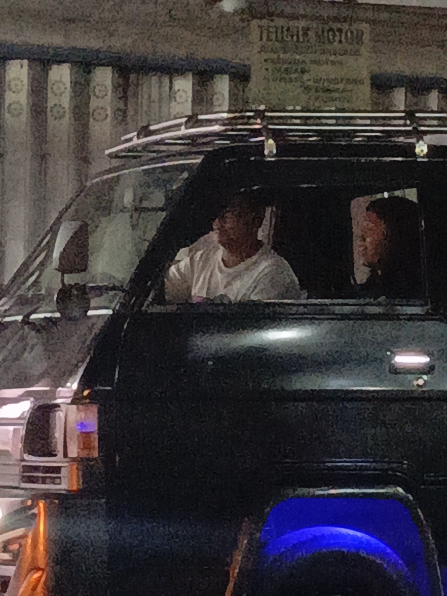 In Purwoharjo, Indonesia, a dark-colored van with a roof rack is parked outdoors at night. The van's windows are tinted. Inside the driver's seat, a person wearing a white t-shirt is visible. In the passenger seat, another person with dark hair is seated. The background shows a building with vertical striped patterns and a sign with the words "TEUFEL MOTOR" and smaller text. The scene is illuminated by ambient streetlights and internal vehicle lights, including a blue light emanating from the front of the van.