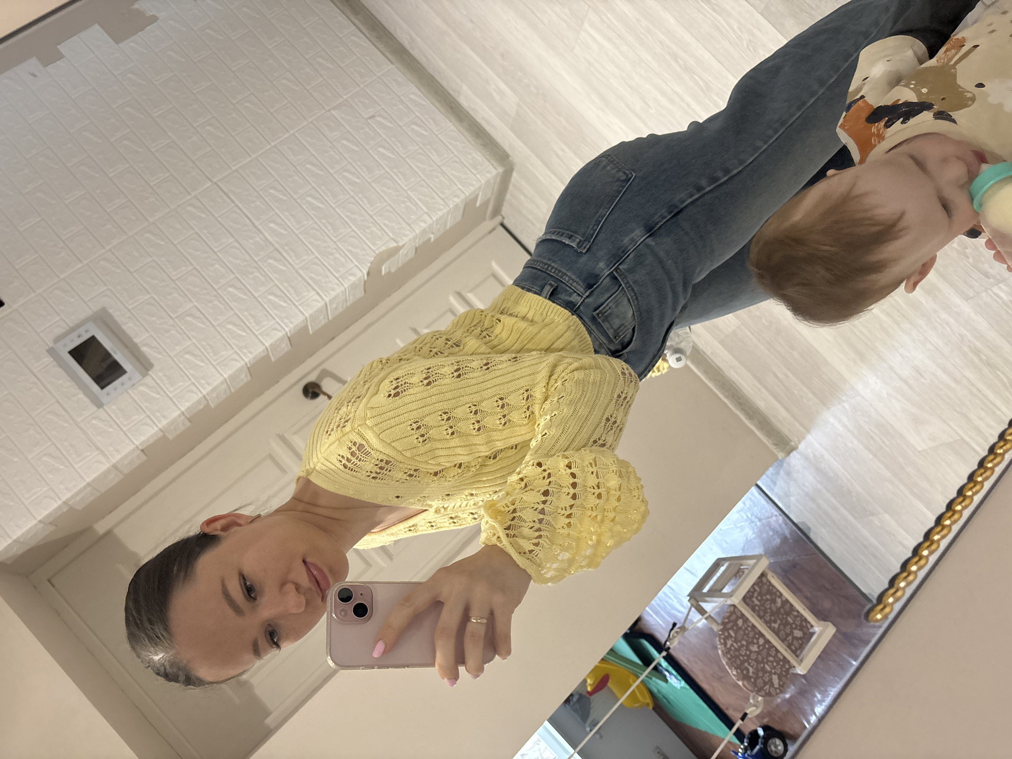 The image displays a mirror selfie of a woman and a baby, captured in an indoor setting. The scene is oriented sideways, with the reflections providing the main view.
The woman, positioned on the left, is holding a light pink smartphone, using it to take the picture. She has dark hair styled away from her face and wears a light yellow, open-knit long-sleeved top and blue jeans. Her fingernails are painted with light pink polish, and she shows a gentle smile.
To her right, a baby or toddler is visible, consuming liquid from a light green bottle. The baby has light brown hair and is dressed in a beige top featuring a brown and orange abstract pattern, seemingly focused on drinking.
The background elements, also reflected in the mirror, show details of a room. A white wall with a distinct raised brick-like texture is seen on the upper left, where a white rectangular panel with a dark screen (possibly an intercom or control unit) is mounted. The floor appears to be light-colored laminate or wood. In the lower right corner of the reflection, hints of children's items are visible, including a small wooden structure resembling a toy or play furniture, and part of a blue toy. A decorative gold chain or trim lines the right edge of the mirror.
The overall impression is that of a candid, domestic moment shared between a mother and child in their home. The specific location context is Ферма, Russia. No visible text is discernible in the image.