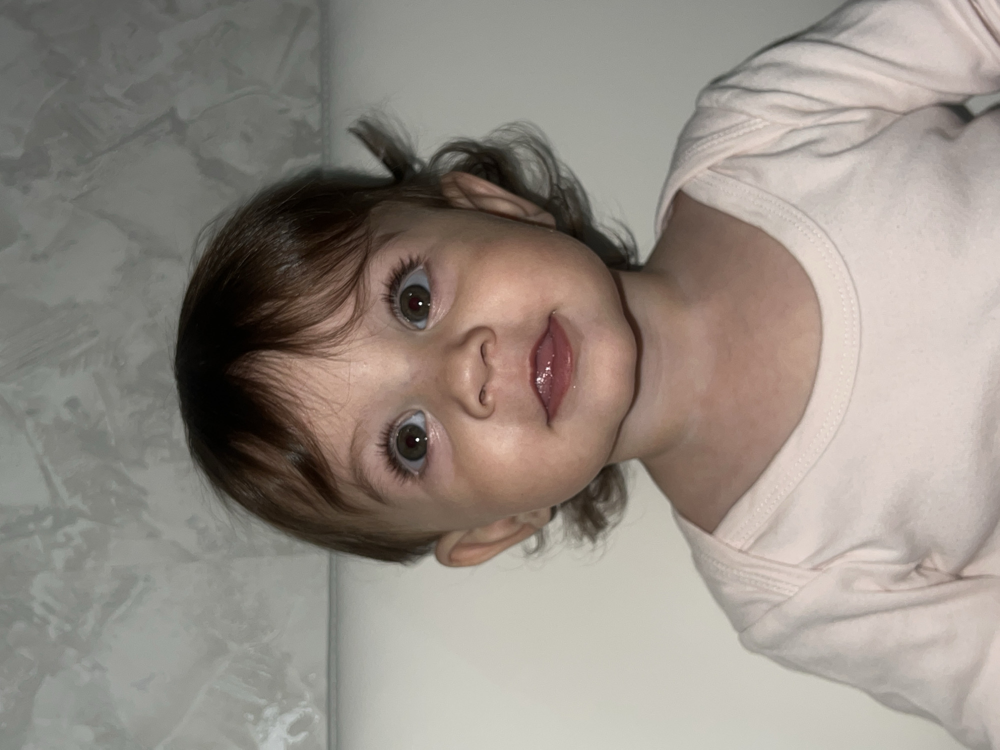 A young child with brown hair and large, light-colored eyes is looking directly at the camera with a slight smile. The child is wearing a light pink, long-sleeved garment. The background appears to be a plain wall with a textured area that could be a pattern or discoloration. The photograph is rotated, placing the child on a diagonal axis. The scene is indoors. It cannot be determined that this image has any association with the city of Elena, Bulgaria.