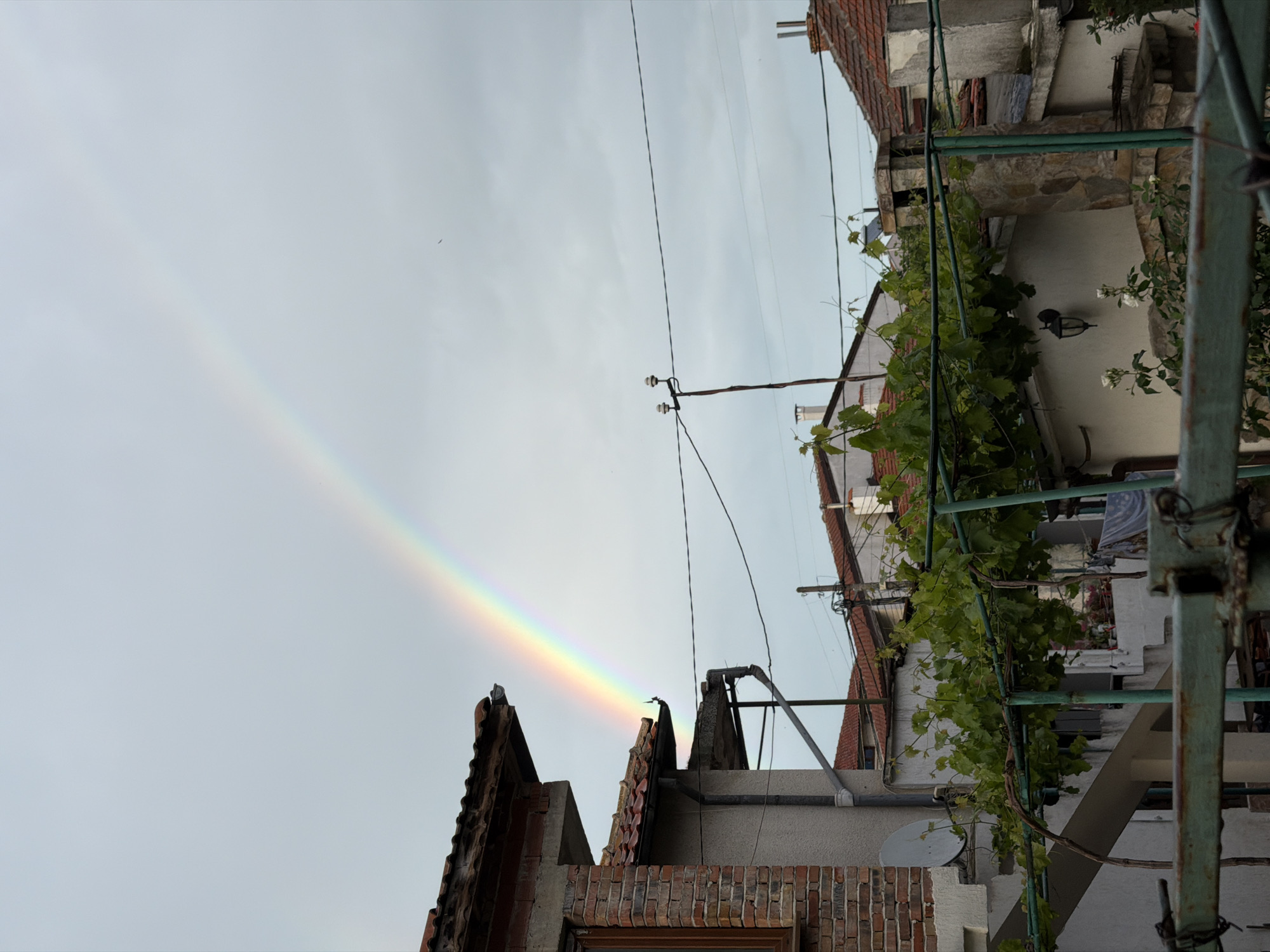 The image shows a partial rainbow ending behind a building in Burgas, Bulgaria. The building has a brick exterior and a tiled roof. There are electrical wires and a satellite dish visible. Vegetation is growing on the side of an adjacent building. A rusty metal structure is in the foreground. The sky is overcast.