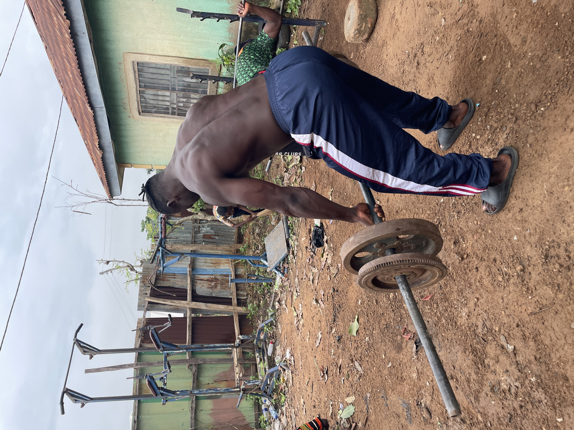 A man performs a lever row on a weighted barbell in Akwanga, Nigeria. The man is shirtless and wearing dark blue track pants with white stripes and gray slides. The weight consists of two rusted, circular metal plates. The environment is a dirt ground. In the background, there is a building with a rusted metal roof, and a window. A homemade weight training setup is also in the background.