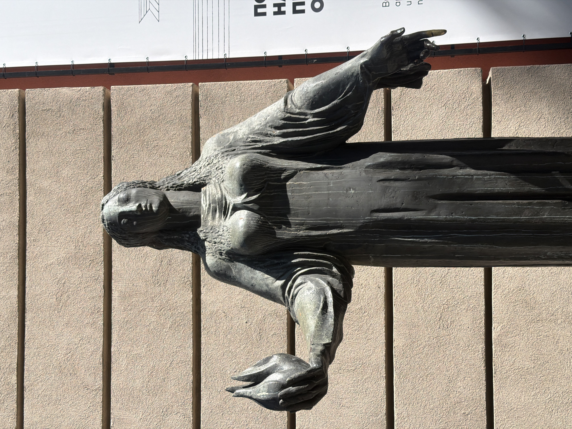 A bronze statue of a woman is fixed to the exterior wall of a building. The statue is in a horizontal position, with the woman's head to the left and her arm extending to the right with her finger pointing. The building's wall is composed of vertical tan-colored concrete panels. At the top of the frame, a partially visible sign is mounted on the wall above the statue. The location of the image is in Sofia, Bulgaria.