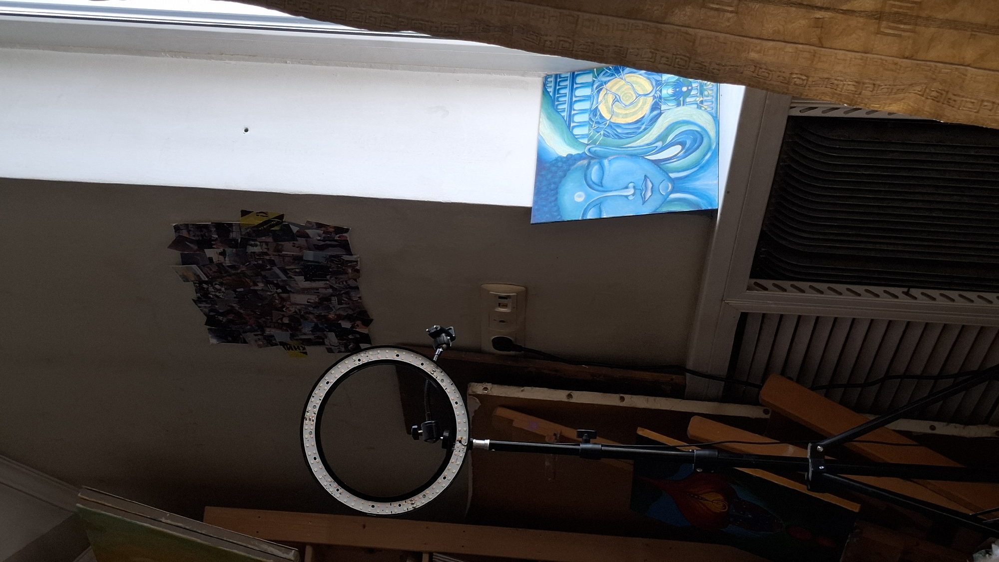This image captures an interior scene in Novaya Vasil'evka, Russia, depicting a room corner illuminated by natural light entering from a window on the right. A prominent black ring light on a tripod stand is positioned on the left, its LED array unlit. Behind the ring light, various canvases or artboards are stacked against a wooden shelf unit. On the wall above the ring light, a dense, irregularly shaped collage of numerous small photographs or printouts is affixed. An electrical outlet with two sockets, into which a black power cord is plugged, is visible on the wall to the right of the ring light.

To the right, a large white window frame or wall section is brightly lit, displaying a small dark mark approximately centered vertically. A vibrant blue painting, depicting a stylized human face with closed eyes, intricate swirling patterns, architectural arches, and a circular yellow luminous form, rests on the windowsill or a ledge below the window. Below this ledge, a heating unit or radiator with vertical fins is partially visible, obscured by a louvered panel. A patterned golden curtain or drape hangs vertically on the far right, partially framing the window opening. The room's overall ambiance is subdued, with brighter illumination concentrated near the window.