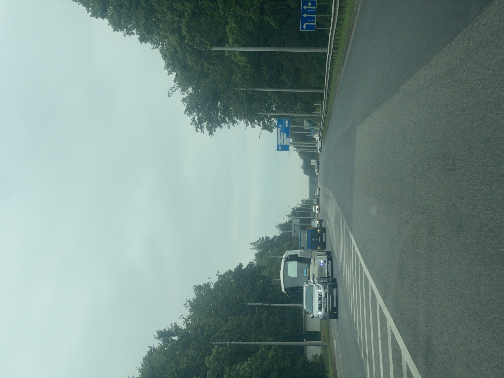 The image shows a multi-lane road with trees lining both sides. The sky is overcast. In the foreground, the road is asphalt with white lane markings. On the left side of the road, a silver SUV is driving in the right lane. Behind it, a white bus is also driving in the right lane. Further ahead, a blue truck is in the right lane. On the right side of the road, there are multiple traffic signs indicating lane directions. The environment appears to be a suburban or semi-rural area. The location is identified as Kopyli, Ukraine.